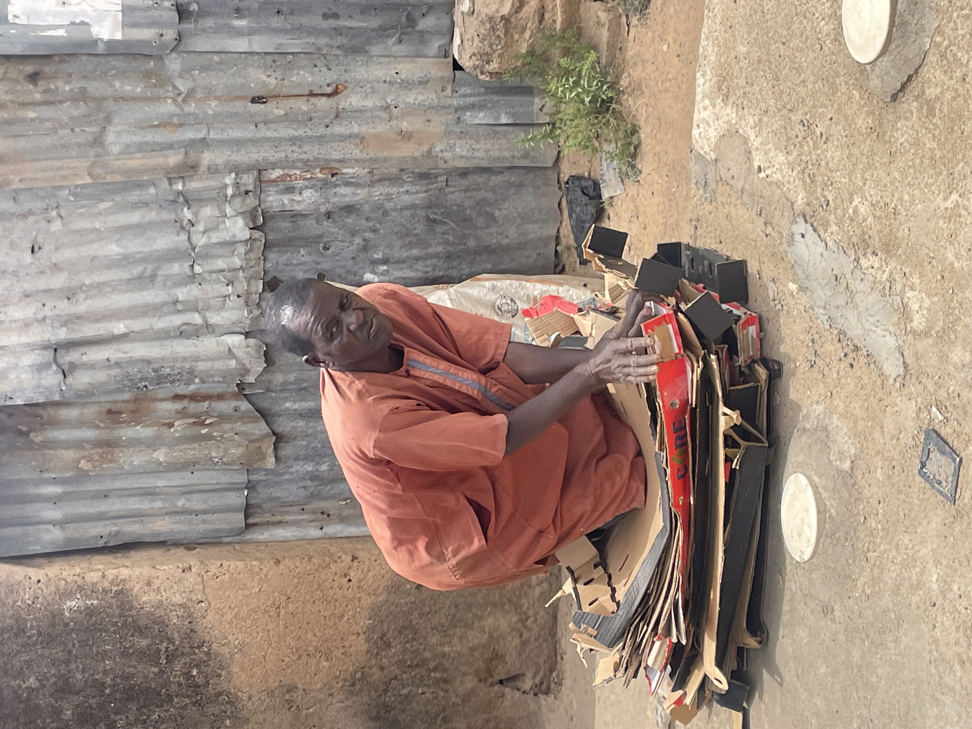 In Jos, Nigeria, an elderly man in an orange outfit is sorting through a large pile of cardboard boxes. He is positioned near a corrugated metal wall and a rough, concrete-like ground surface. The boxes appear to be from various sources. The scene suggests an informal recycling or scavenging activity.