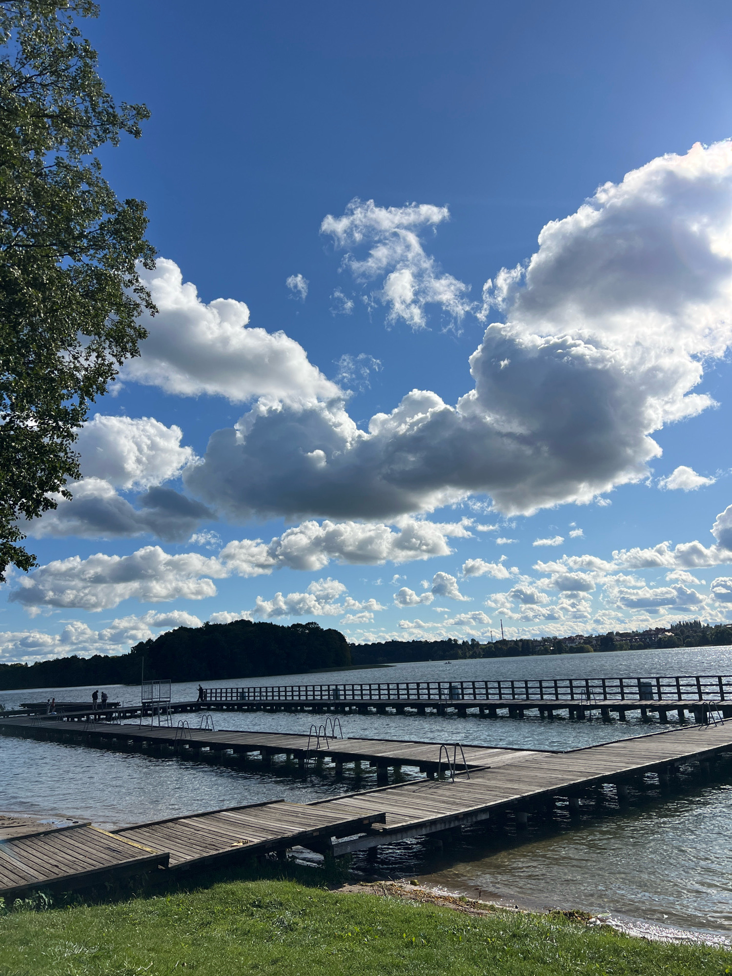 The image depicts a wide, calm lake with a clear blue sky filled with fluffy white clouds. Several wooden piers extend into the water at various angles, some with railings and ladders. A shoreline with green grass is visible in the foreground. In the distance, a tree-covered island and a town with buildings can be seen. A few people are visible on the piers. The overall scene suggests a recreational area on a sunny day. This scene is located in Mrągowo, Poland, a town known for its many lakes.