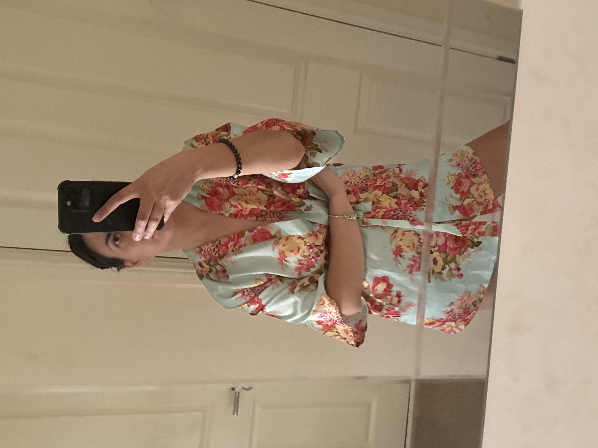 A woman is taking a mirror selfie in what appears to be a home interior. She is wearing a light blue satin robe with a floral print of pink and yellow flowers. The robe has wide sleeves and is cinched at the waist. She has dark hair pulled back and is holding a black smartphone in her right hand, obscuring part of her face. On her left wrist, she wears a dark beaded bracelet, and on her right wrist, a silver bracelet with a charm. She is standing with her arms crossed. The background shows a plain, light-colored wall and a door with a silver handle. The floor in the foreground is a reflective surface, likely a dresser or table, showing a reflection of the lower part of the robe and the woman's legs. The scene is well-lit, suggesting indoor lighting. There is no indication of the image being located in Great Yarmouth, United Kingdom, or any specific city.