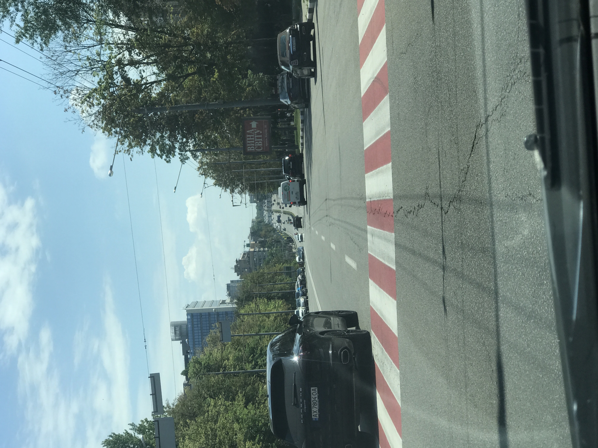 The image shows a street scene in a city. The perspective is from inside a vehicle, looking forward. In the foreground, a crosswalk with red and white stripes is visible on the asphalt road. A black Porsche Cayenne is in the left lane, stopped at the crosswalk. Its license plate reads AX 7804 OA. Other cars are further down the road in various lanes. Trees line both sides of the street. In the background, several multi-story buildings are visible, including a modern glass building. The sky is blue with scattered white clouds. A sign in Ukrainian is visible on the right side of the road. The overall impression is a moderately busy urban street on a sunny day.
