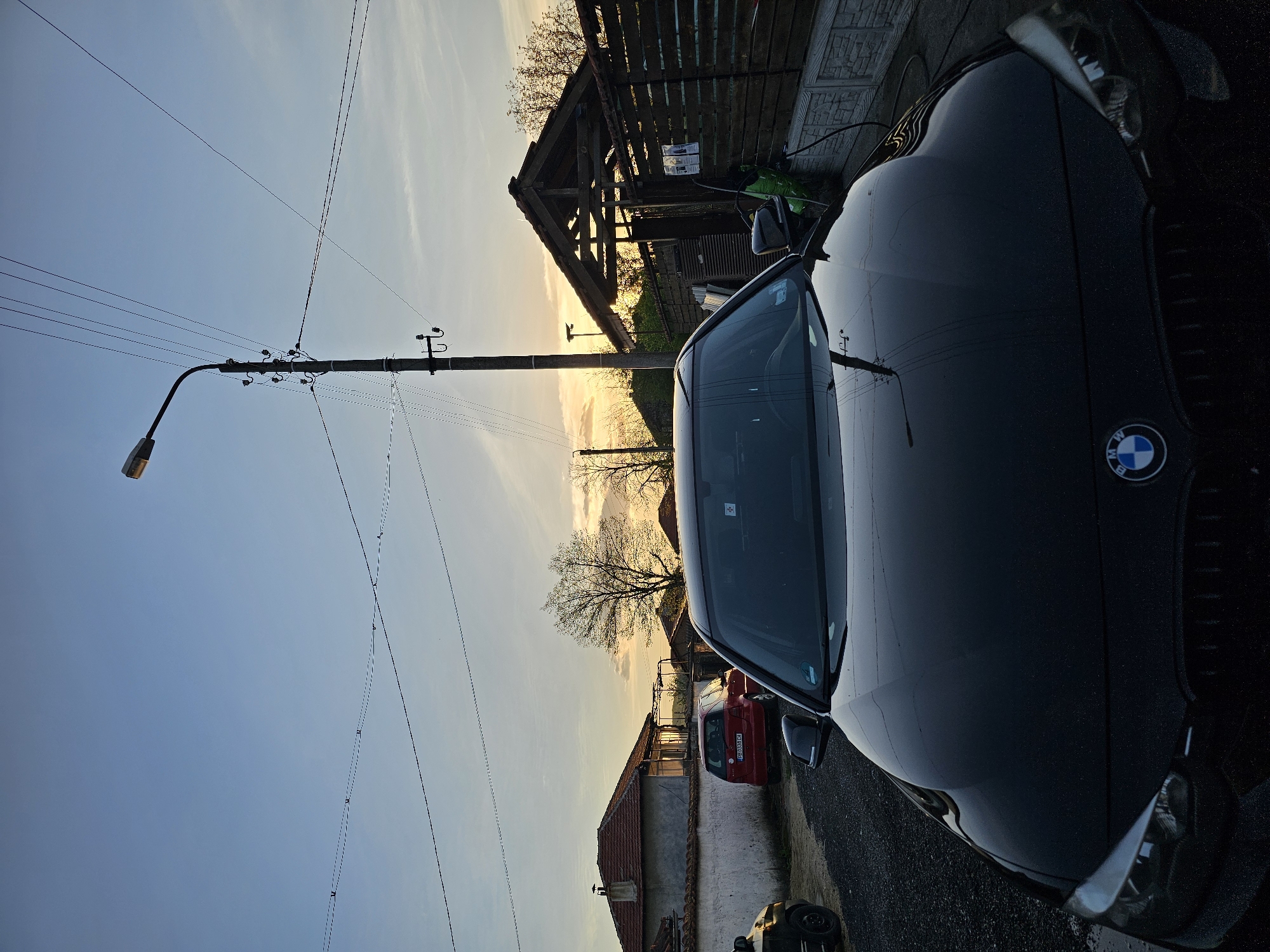 The image captures a tranquil street scene at either sunrise or sunset, characterized by a warm, golden glow on the horizon and a clear, blue sky overhead. The setting is a residential street, likely in a rural or suburban area of Krasnovo, Bulgaria.
In the foreground, a dark-colored BMW SUV is parked on the right, its shiny hood and windshield reflecting the surrounding sky and a nearby utility pole. The distinctive BMW logo is clearly visible on the vehicle's grille. To the left, a tall, black utility pole equipped with a street light and numerous electrical wires stands prominently, with wires stretching across the sky.
Mid-ground details include an asphalt road surface that appears slightly reflective. Further down the street, a red hatchback car is parked, and its Bulgarian license plate, "EB 0894 CA," is partially visible. In the background, various buildings are seen, including a wooden structure with a sloped roof behind the BMW, and a house with a red tiled roof further down the street. Bare-branched trees silhouette against the bright horizon. There is no discernible activity or any people present, contributing to a serene atmosphere.