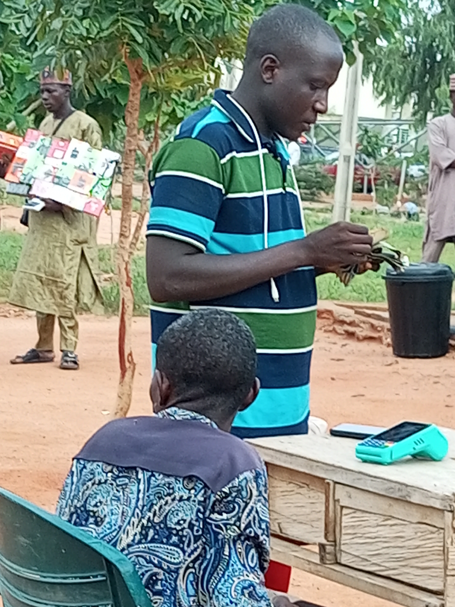 In Gombe, Nigeria, on a dusty outdoor market ground, a man wearing a blue, green, and white striped shirt is counting money. To his left, another man in a long, olive-colored robe and a patterned cap is holding a stack of colorful items, possibly soap or food products. In the foreground, a person wearing a blue and white patterned shirt is seated in a green chair, facing away from the camera. A wooden table is positioned between the seated person and the man counting money. On the table, a light blue mobile payment terminal and a smartphone are visible. A black bucket is located near the man counting money. The environment is characterized by dry, sandy ground and sparse green foliage.