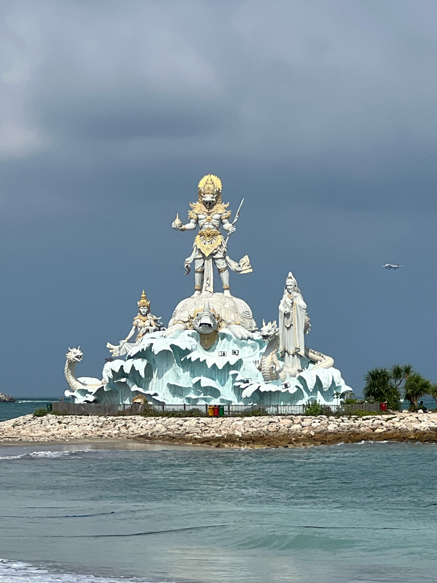The image shows the Garudha Wisnu Kencana Cultural Park in Kuta, Indonesia. The park features a large statue of the Hindu deity Vishnu riding on the mythical bird Garuda. The statue is surrounded by other sculptures, including a dragon and a turtle. The park is located on a hill overlooking the ocean, and in the background, a plane is visible in the sky. The sky is overcast, and the ocean is a deep blue.