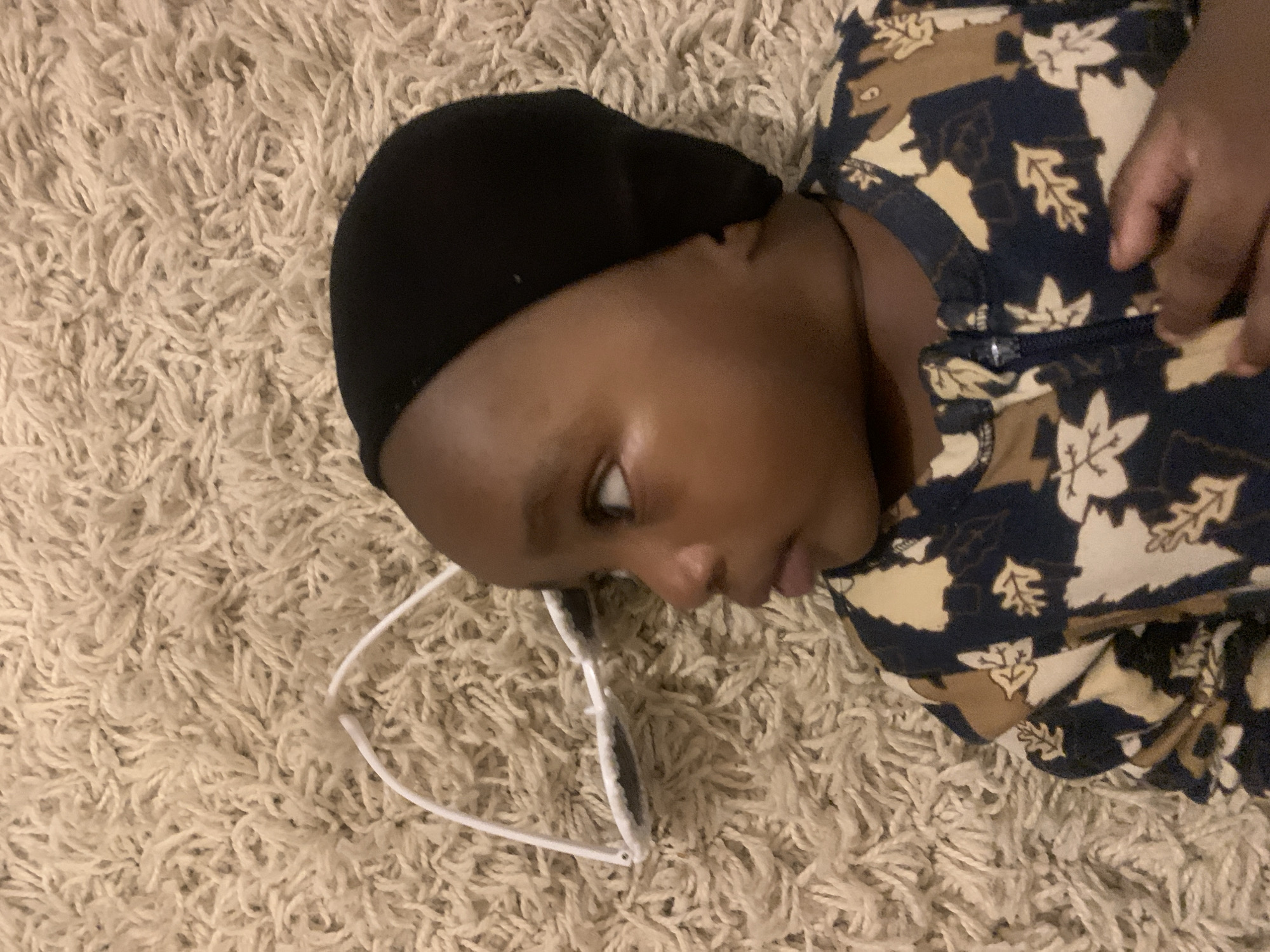 A young child lies on a light-colored shag carpet, wearing a black cap and a dark floral-patterned top. White-framed glasses rest on the carpet near the child's head. The child is looking to the side. The location is not discernible as Barakin Akawo, Nigeria from the visual information provided.