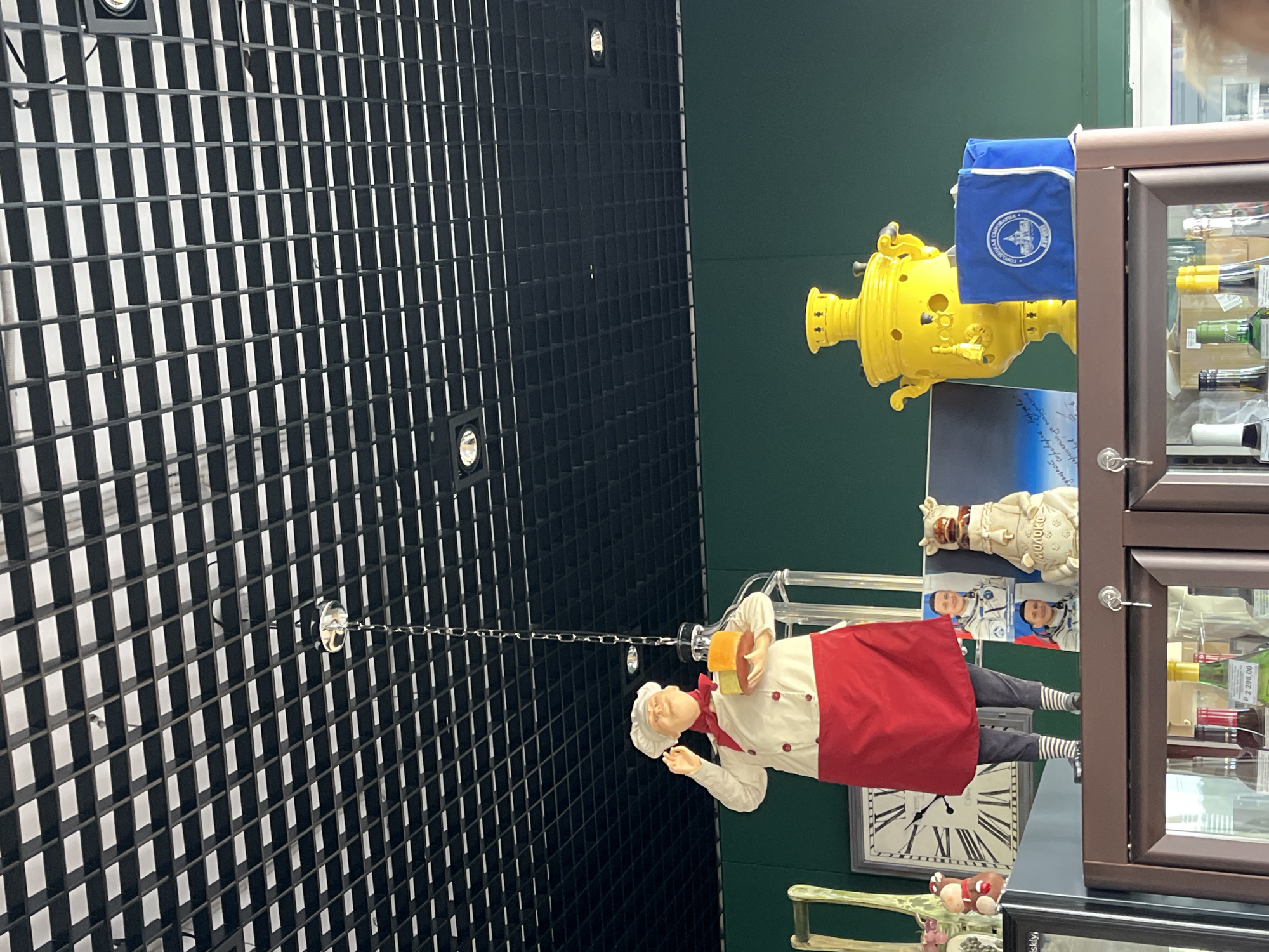 This image captures a display on a shelf, likely in a shop or restaurant, in the village of Akhlebaikha (Kovriginskii S/S), Russia. The backdrop is a dark green wall. Dominating the left side of the display is a life-sized mannequin of a chef, wearing a white chef's coat, a red apron, and a red neckerchief. The chef holds a transparent container with what appears to be cheese or food samples. To the right of the chef, a large, bright yellow samovar is prominently displayed. Next to the samovar is a blue fabric bag with a circular white logo featuring a building and text. Behind the chef, a large wall clock with Roman numerals is visible. To the right of the clock and slightly in front of it are two framed pictures of men in astronaut suits. A small, white decorative object resembling a cow or bull, adorned with a bell, is positioned between the astronaut pictures and the samovar. On the shelf below, various bottles of alcohol are visible through the glass doors of a cabinet. The ceiling above features a black grid with recessed lighting. The overall scene appears to be a themed display, possibly related to culinary arts or Russian culture, set within a commercial establishment.