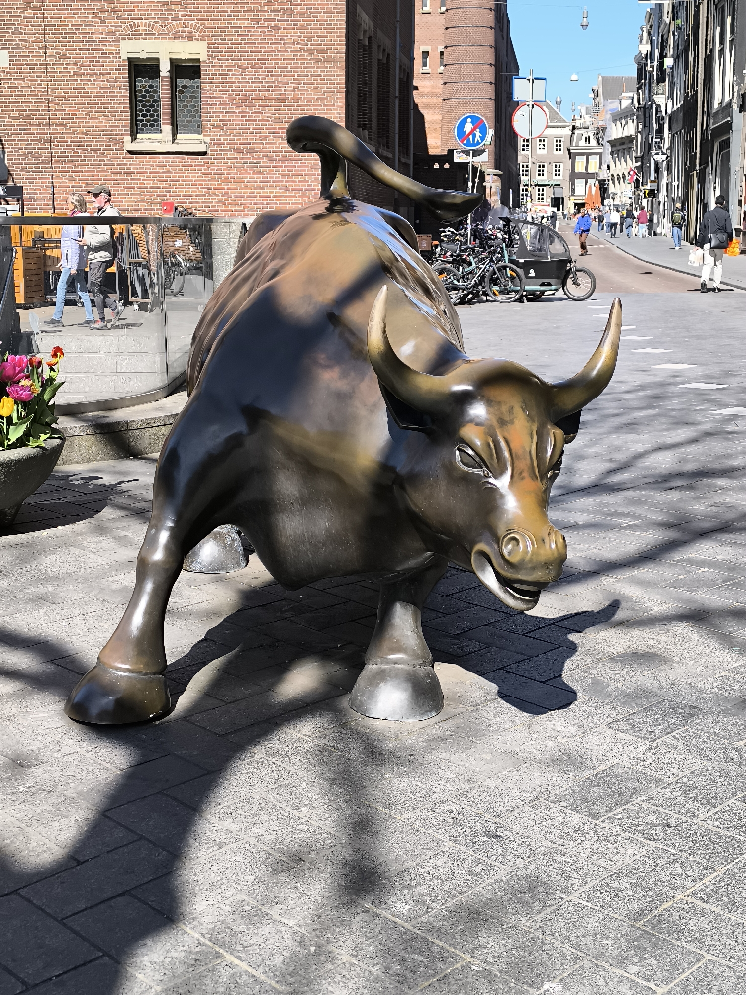 A bronze bull statue stands prominently on a paved area in Amsterdam, Netherlands. The bull is the central subject, its form casting shadows on the pavement. In the background, pedestrians are visible walking along the street, along with bicycles parked near a building. The street scene includes buildings with typical Dutch architecture and signage. A planter with tulips is situated in the lower left corner. The overall image depicts a sunny urban scene with the bull statue as a notable feature.