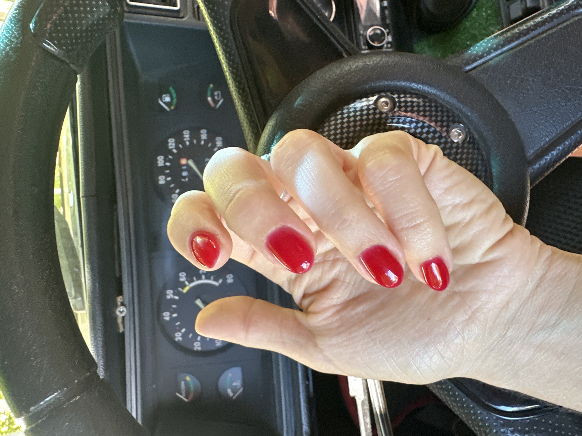 A close-up shot shows a person's left hand with red-painted fingernails inside a car, positioned in front of a steering wheel and dashboard. The hand is resting on the steering wheel, which has a textured grip. The dashboard features various gauges, including a speedometer. The scene appears to be in a city, possibly Novoaltaysk, Russia, based on the provided location information.