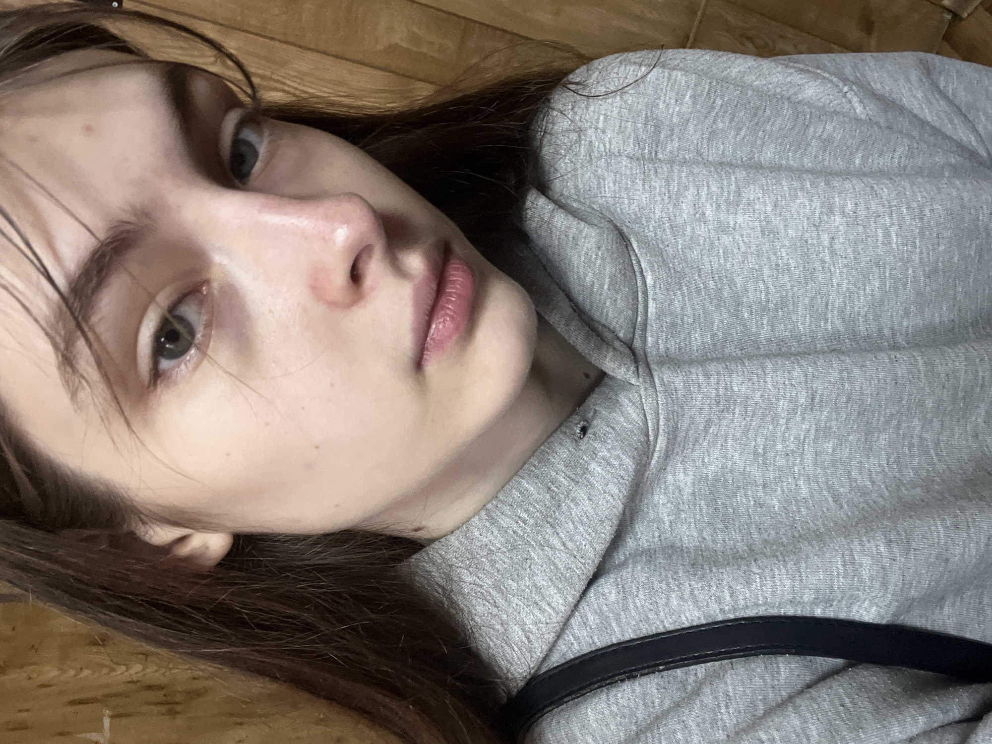 A young woman lies on a wooden floor, her head resting near the bottom left corner of the frame. She is wearing a grey hooded sweatshirt, and a black strap is visible across her torso. Her face is angled towards the camera, with her brown hair partially obscuring the floor beneath. Her complexion is fair, and her gaze is directed towards the upper left. The background is a simple wooden floor with a warm tone. There is no information available within the image to determine the location as Saint Petersburg, Russia.