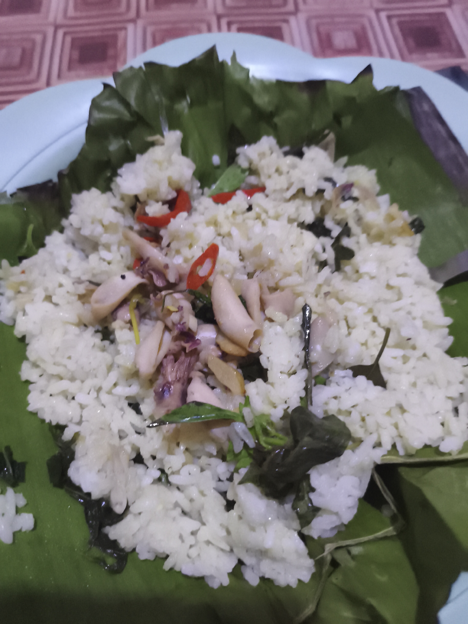 A close-up, overhead shot features a plate of white rice served with squid and vegetables on a banana leaf. The rice is fluffy and mixed with pieces of squid, red chili peppers, and green herbs. The banana leaf is neatly folded around the rice, creating a natural serving dish. The plate is light blue, and the background shows a brown and cream patterned tablecloth. The food appears to be a home-cooked meal, possibly prepared and consumed in a residential setting in Depok, Indonesia. There are no people visible in the image, and the focus is solely on the food.