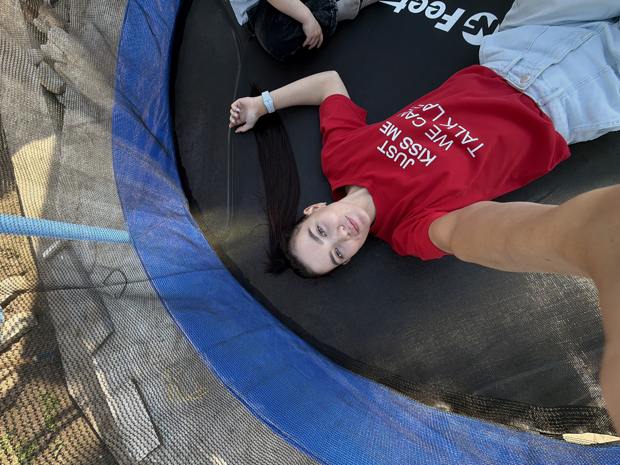 The image is a selfie taken from above, showing a woman lying on a trampoline. She has dark hair, fair skin, and is wearing a red t-shirt with the phrase "JUST KISS ME WE CAN TALK LATER" printed in white. She is also wearing light-colored jeans and a light blue wristband. Another person, presumably a child, is partially visible in the upper left corner, wearing dark clothing and grey socks. The trampoline has a black jumping surface and a blue padded edge. A safety net surrounds the trampoline. The location appears to be outdoors, possibly in a backyard or garden. While it's impossible to pinpoint the exact location in Chaikovsky, Russia, based solely on this image, it's suggested it's a residential area.