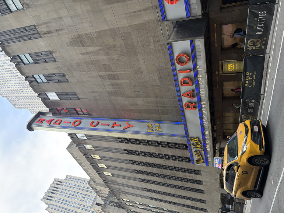 The image displays the exterior of Radio City Music Hall, located in Midtown Manhattan, New York City, United States. The building presents an Art Deco architectural style with a light gray stone facade featuring prominent vertical fluting and recessed windows. A tall, vertical sign on the building's left side reads "RADIO CITY" in red neon with blue trim, with "Music Hall" displayed below in a smaller vertical format.
A marquee extends over the sidewalk, prominently featuring "RADIO CITY" in red neon letters outlined in blue, above "Music Hall". Below this, event information is displayed in white text against a dark background, stating "RAYE APR 15 & 16" and "ZAKIR KHAN FRI APR 17".
On the street in the foreground, a yellow New York City taxi, specifically a Toyota Sienna minivan, is driving from left to right. The sidewalk directly in front of the building is secured by black temporary barricades. These barricades bear the "RADIO CITY MUSIC HALL" logo and "100 YEARS OF ROCKETTES 1925-2025" branding. Several individuals are visible on the sidewalk near the building's entrance doors, with some appearing to walk or stand. In the background, taller skyscrapers are visible against an overcast sky.