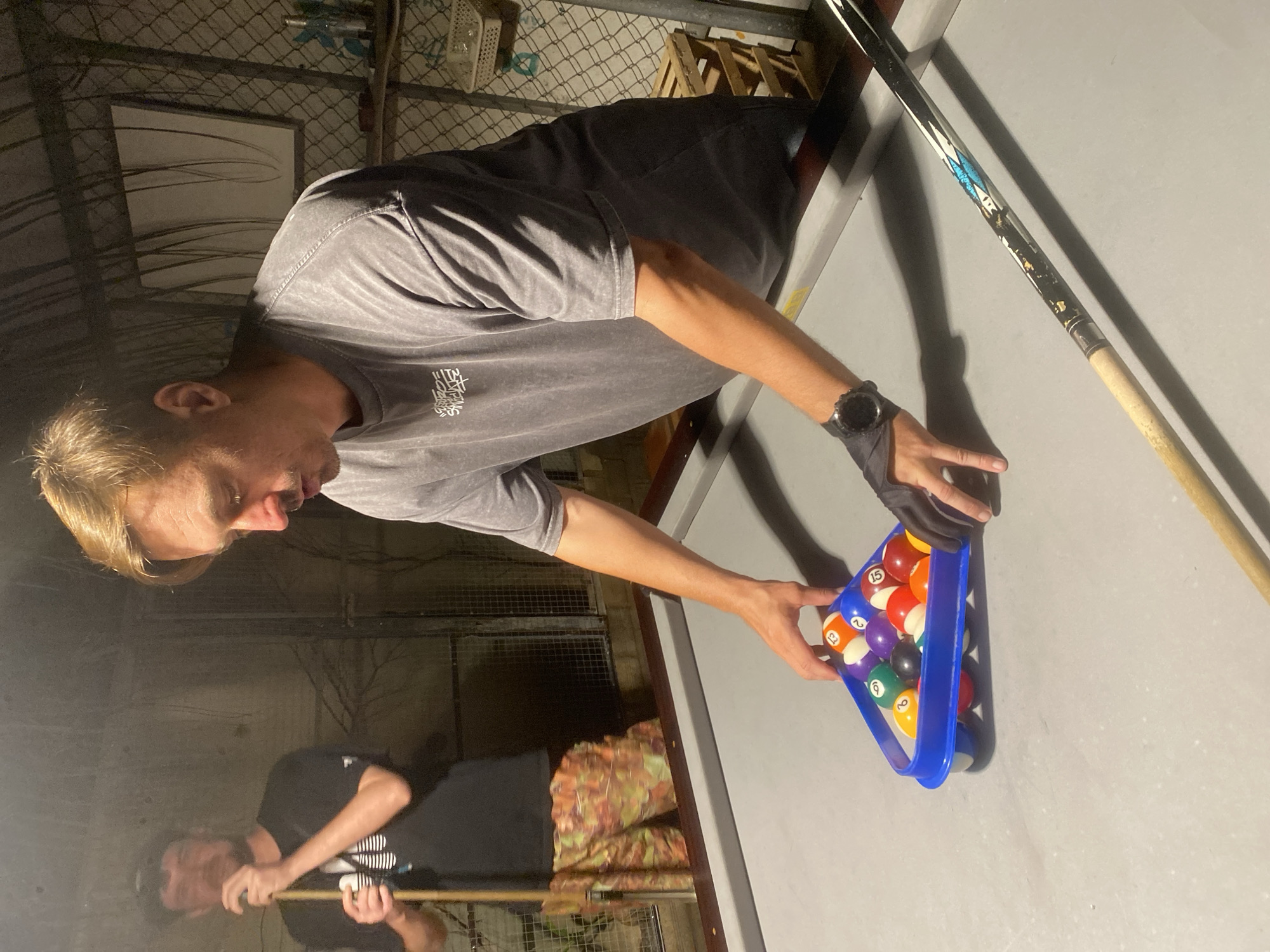 A man is setting up a rack of billiard balls on a pool table. He wears a gray t-shirt, shorts, and a watch. He holds a blue rack filled with pool balls. A pool cue rests on the table next to the rack. Another person is visible in the background holding a cue. The setting appears to be in a covered outdoor area. The photograph was taken at night in Mũi Né, Vietnam.