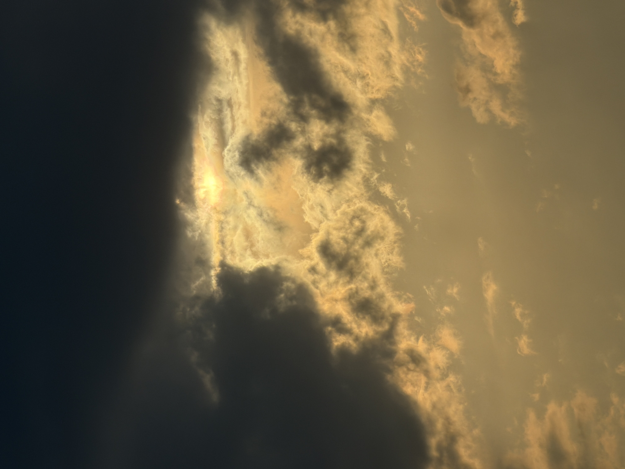 The image shows a cloudy sky, with the left side dominated by a large, dark cloud formation. Through a break in the clouds, a bright, golden light shines, suggesting the sun is behind the clouds. The rest of the sky has scattered clouds, also tinged with the same golden light. The image is rotated 90 degrees clockwise.
It is not possible to determine the location in the city of Tbilisi, Georgia from the image.