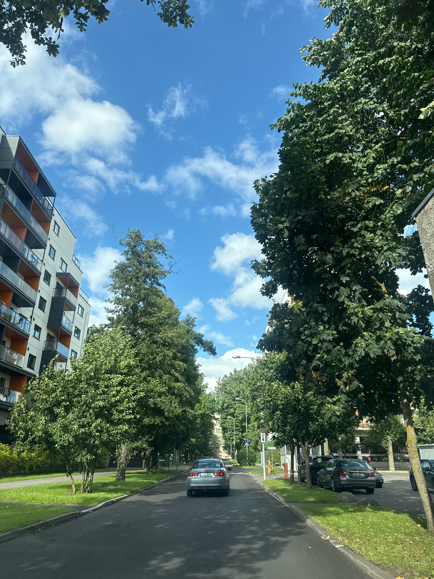 The image shows a street scene in Tallinn, Estonia, on a sunny day with a blue sky and scattered white clouds. On the left side of the street is a modern apartment building with balconies. On the right side, there are several trees lining the sidewalk, and parked cars. In the foreground, a silver sedan is driving away from the camera. The road is paved, and there is a grassy area between the road and the sidewalk. Further down the street, more trees and buildings are visible.