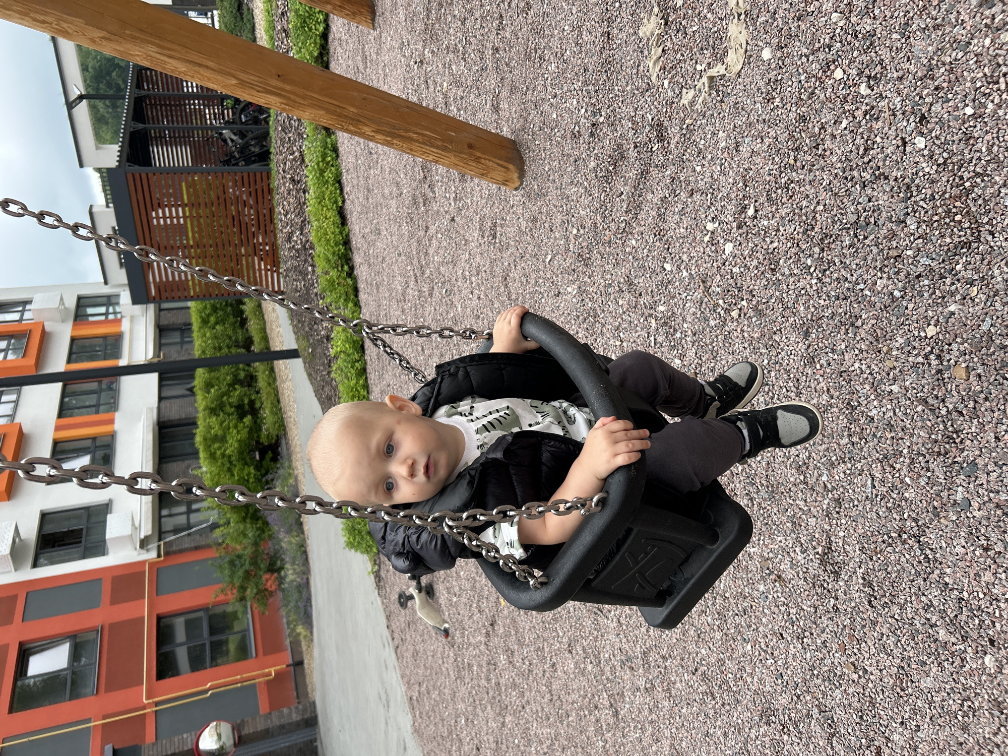 A young Caucasian male child, appearing to be around one to two years old, is seated in a black plastic baby swing. He is wearing a black puffer vest over a white t-shirt with a green and black abstract pattern, dark gray pants, and black and gray sneakers. The child has blond hair and blue eyes and is looking to his right. The swing is suspended by metal chains from a wooden swing set, the right support of which is visible in the foreground. The ground beneath the swing set is covered in small, multi-colored gravel. In the background, there is a modern apartment building with orange, white, and gray facades, and a covered bicycle parking area with wooden slats. The location is identified as a playground in the city of Kaluga, Russia.