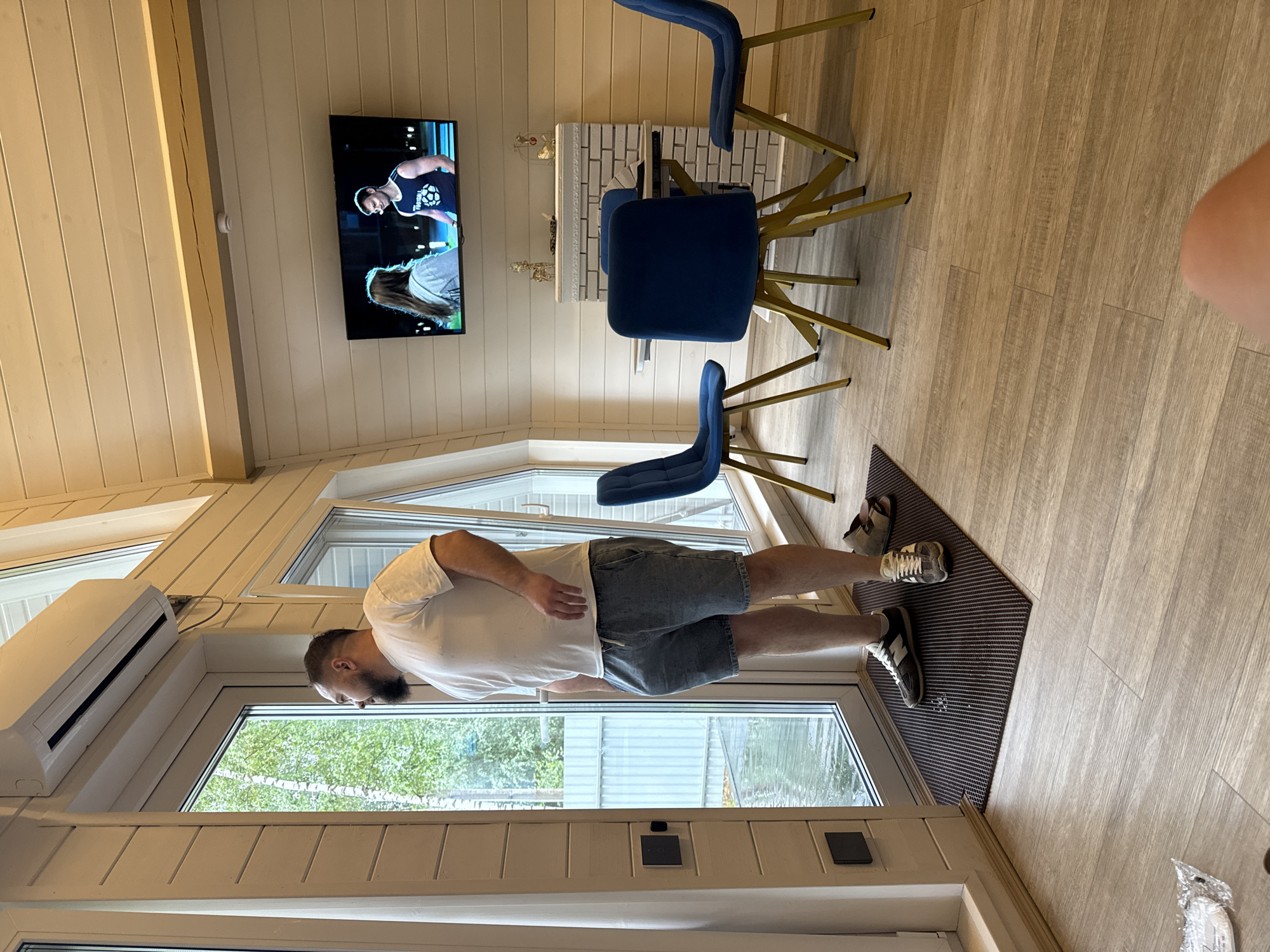 The image depicts an interior scene, likely a room with wooden paneled walls and a sloped ceiling. In the foreground, a man stands near a large glass door, looking outwards. He is wearing a white t-shirt and denim shorts, with sneakers on his feet. A doormat is positioned by the door, and a pair of sandals rests on it.
To the right of the man, a small dining area is set up with a round table and three blue chairs. Above the table, a television is mounted on the wall, displaying an image of a man and a woman. A decorative fireplace with a white brick facade is visible behind the table.
An air conditioning unit is mounted high on the wall near the glass door. The room appears to be well-lit, with natural light entering through the door and windows. The flooring is a light-colored wood laminate. The outdoor scenery visible through the glass suggests a natural environment with trees. The image does not contain any specific visual cues that would allow for its location to be identified as Prokudino, Russia.