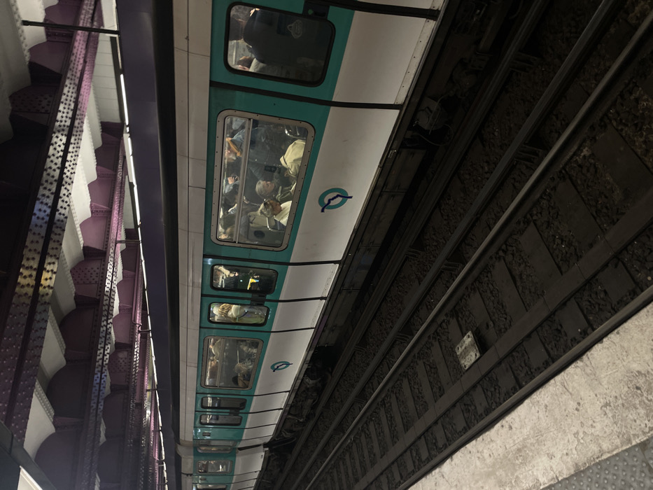 A diagonally-angled shot shows a Paris Metro train at a station. The train, white with a teal stripe, is filled with passengers visible through the windows. To the left is a station support structure with purple arches. To the right are the train tracks, composed of metal rails and gravel. A concrete platform edge is visible below the tracks. The lighting is dim and industrial, emphasizing the urban transit environment.