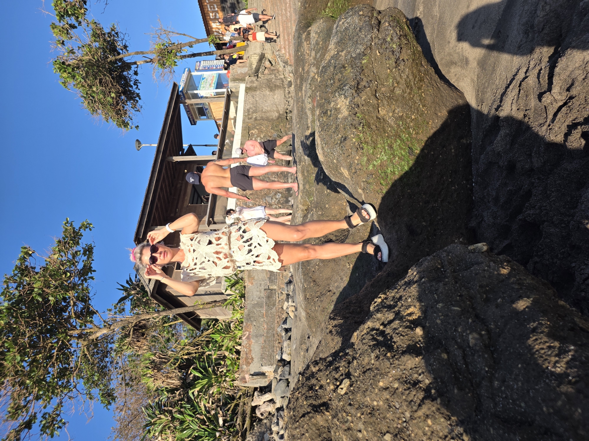 A woman with pink hair is standing on a rock formation at Batu Bolong Beach in North Kuta, Indonesia. She is wearing a light-colored, patterned dress and sandals. She is also wearing sunglasses and is holding them at the top of her forehead. Behind her is a structure with several people underneath and beside it. In the background, there are more people and a blue sky with a few trees. To the right, there is a large, dark rock formation covered with moss. The time of day appears to be early morning or late afternoon.