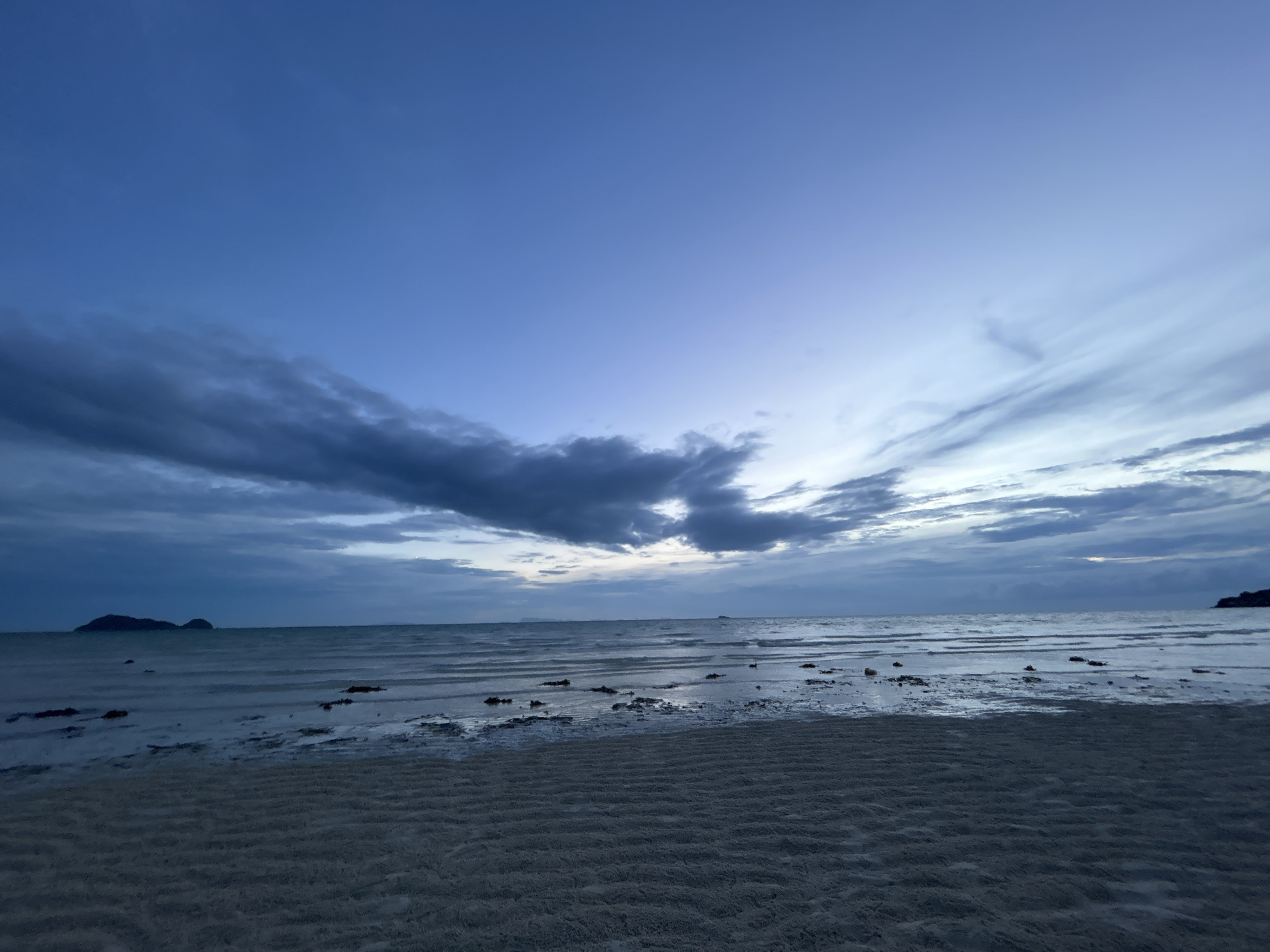 This image captures a serene beach scene at dusk in Ko Pha-ngan District, Thailand. The foreground is dominated by rippled sand, indicating the receding tide. The middle ground features the calm ocean, with shallow water reflecting the twilight sky and scattered dark objects, likely seaweed or rocks, visible in the water. In the distance, a silhouette of islands can be seen on the horizon, adding depth to the seascape. The sky transitions from a deep blue at the top to lighter shades of blue and purple near the horizon, with scattered dark clouds creating dramatic streaks across the sky. The overall mood is peaceful and tranquil, characteristic of a quiet evening by the sea. The time of day is clearly dusk or twilight, indicated by the soft, diffused light and the deep blue hues of the sky. There is no visible text in the image.