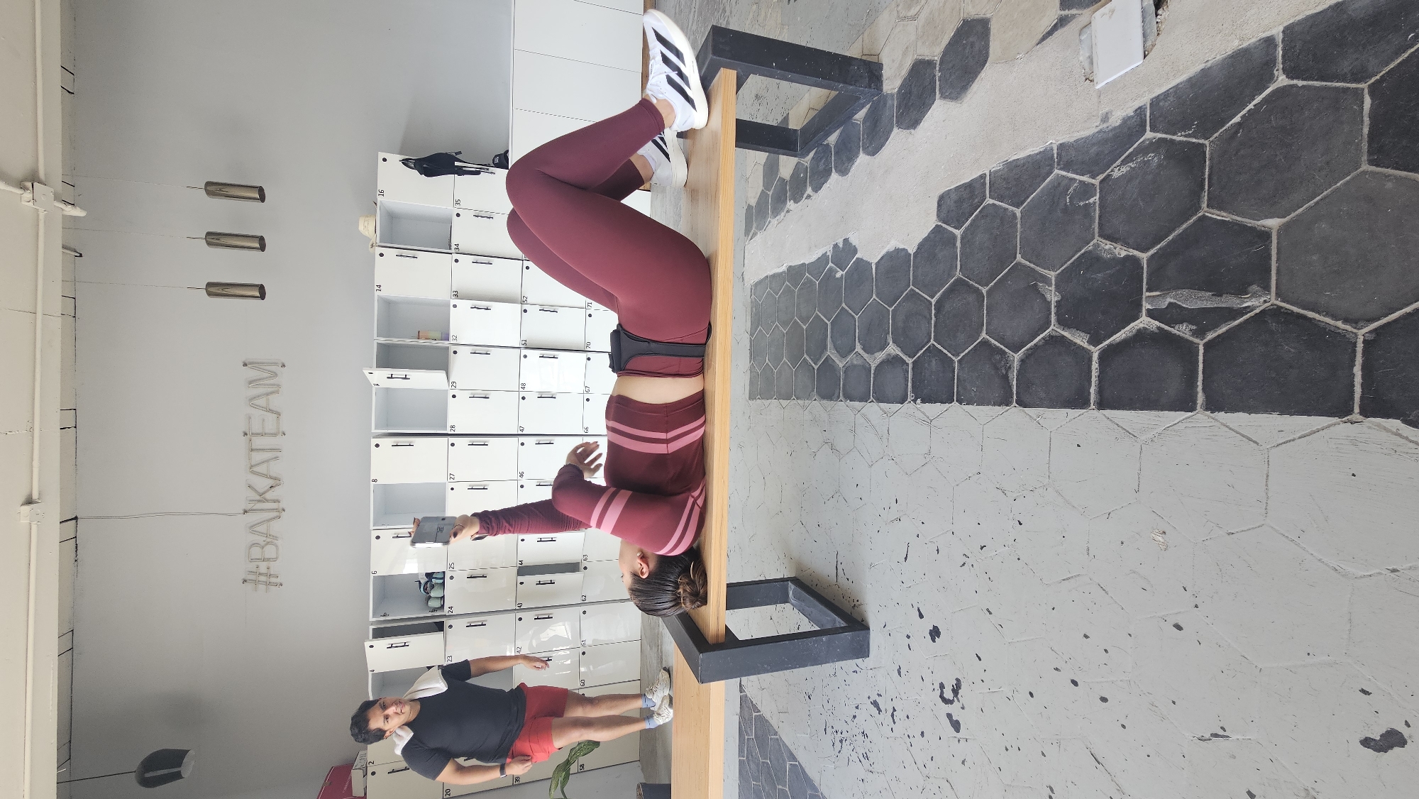 This image depicts an indoor scene within a fitness studio located in Puebla, Mexico, identified by the visible "#BAIKATEAM" neon sign on the back wall.
The central subject is a woman lying on her back on a light-colored wooden bench with black metal legs. She is dressed in a dark maroon athletic outfit, including a long-sleeved crop top with lighter stripes and matching leggings. White Adidas sneakers are visible on her feet. She holds a dark smartphone vertically above her head with both hands, pointing it upwards. A black fitness belt or brace is visible around her waist.
To the left of the bench, partially obscured by a small potted plant, a man stands. He wears a black t-shirt, red shorts, and white sneakers, with a white towel draped over his left shoulder. He is looking towards the woman or the area she is filming.
The background features a long wall lined with numerous white, metal lockers, many of which are numbered (e.g., 23, 24, 25, 27-29, 32-35, 42-48, 51-52, 60-63, 66-71). Several locker doors are open; a black backpack hangs from one on the right. Above the lockers, the wall is light grey, adorned with the prominent white neon sign displaying "#BAIKATEAM." Three cylindrical metallic pendant lights hang from the ceiling.
The floor is comprised of hexagonal tiles, divided into two distinct sections: the left half features light grey tiles with scattered dark splatters, while the right half consists of dark grey tiles, showing some wear. The demarcation between the two tile patterns is a straight line running perpendicular to the bench. The room is brightly lit.