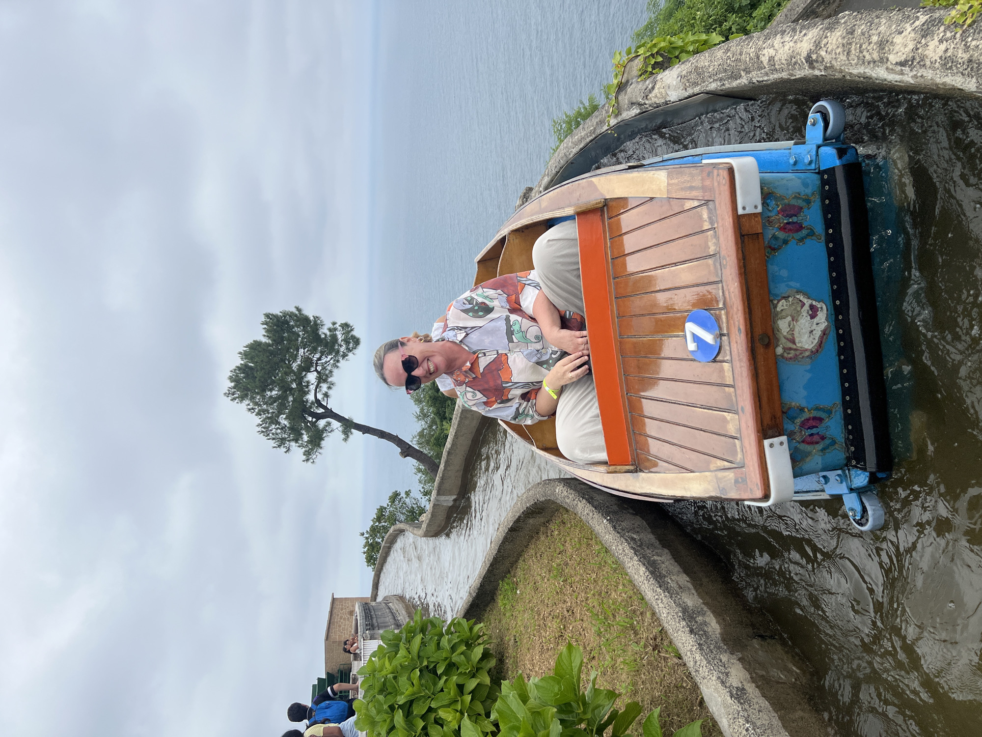 A woman is riding in a wooden boat on a water flume ride. The boat is decorated with blue panels featuring a tiger and a butterfly. A blue circular number "7" is visible on the wooden deck of the boat. The woman is smiling and wearing sunglasses and a colorful shirt. She is seated in the boat with her hands resting on her lap. The water flume follows a winding path alongside a grassy embankment and a concrete wall. In the background, the vast expanse of the sea is visible under a cloudy sky. The location is Monte Igueldo, an amusement park in San Sebastián, Spain, known for its vintage attractions and panoramic views of the Bay of Biscay.