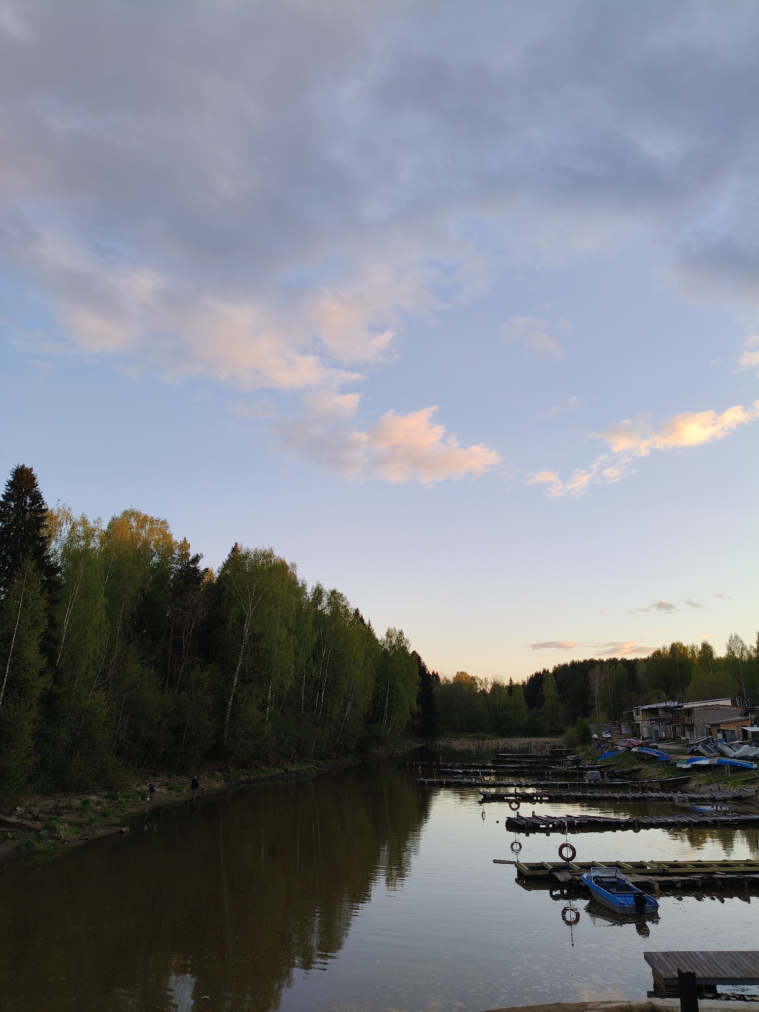 The image depicts a tranquil scene on the Saygatsky Bay in Chaikovsky, Russia. A body of water, presumably a lake or wide river, stretches from the foreground towards the horizon, reflecting the sky and the adjacent treeline. The treeline on the left consists of a mix of deciduous and coniferous trees, some showing signs of new spring foliage. Two figures can be seen on the shore in the lower-left quadrant of the image.
On the right side of the image, several wooden docks jut into the water, with a blue boat moored to one of them. A series of life preservers are attached to the docks.
In the background on the right, several boats and structures are visible, suggesting a boat club or marina. The sky is filled with clouds tinged with the warm colors of the setting sun. The overall lighting suggests it is late afternoon or early evening.