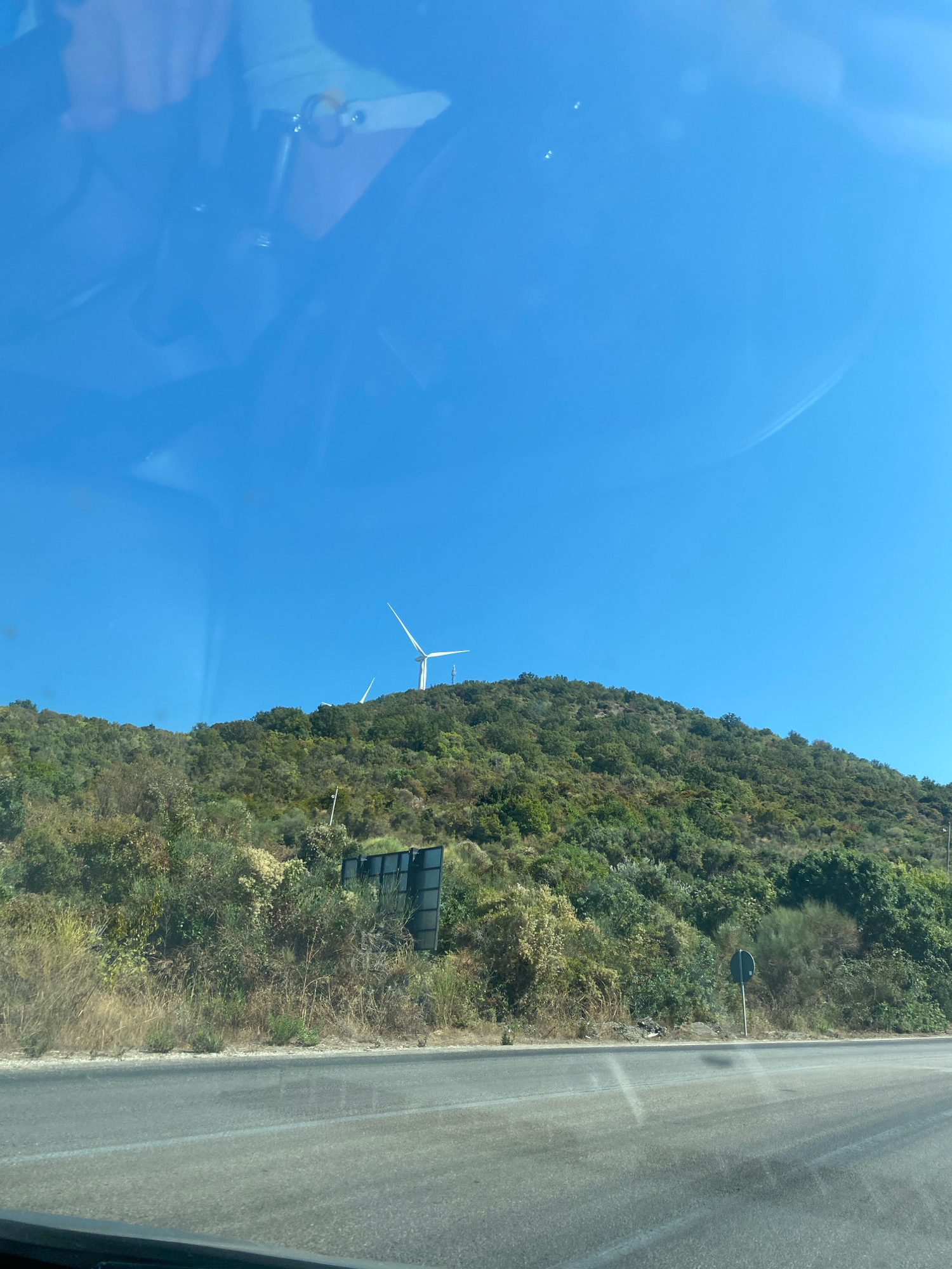The image captures a view from a vehicle, looking out at a treed hillside under a clear blue sky. Two wind turbines are visible on the crest of the hill, with one prominently displayed in the right half of the frame. The hillside is densely covered with a variety of green vegetation, including trees and shrubs, with some drier grasses and bushes along the lower slopes near the road. A dark rectangular structure, possibly a sign or advertising board, is partially obscured by foliage on the hillside. A round road sign on a pole is also visible on the right side of the road. The foreground shows a section of asphalt road with white lane markings. The overall impression is a scenic, rural landscape with renewable energy infrastructure. The location is identified as being in the city of Kunje, Montenegro.