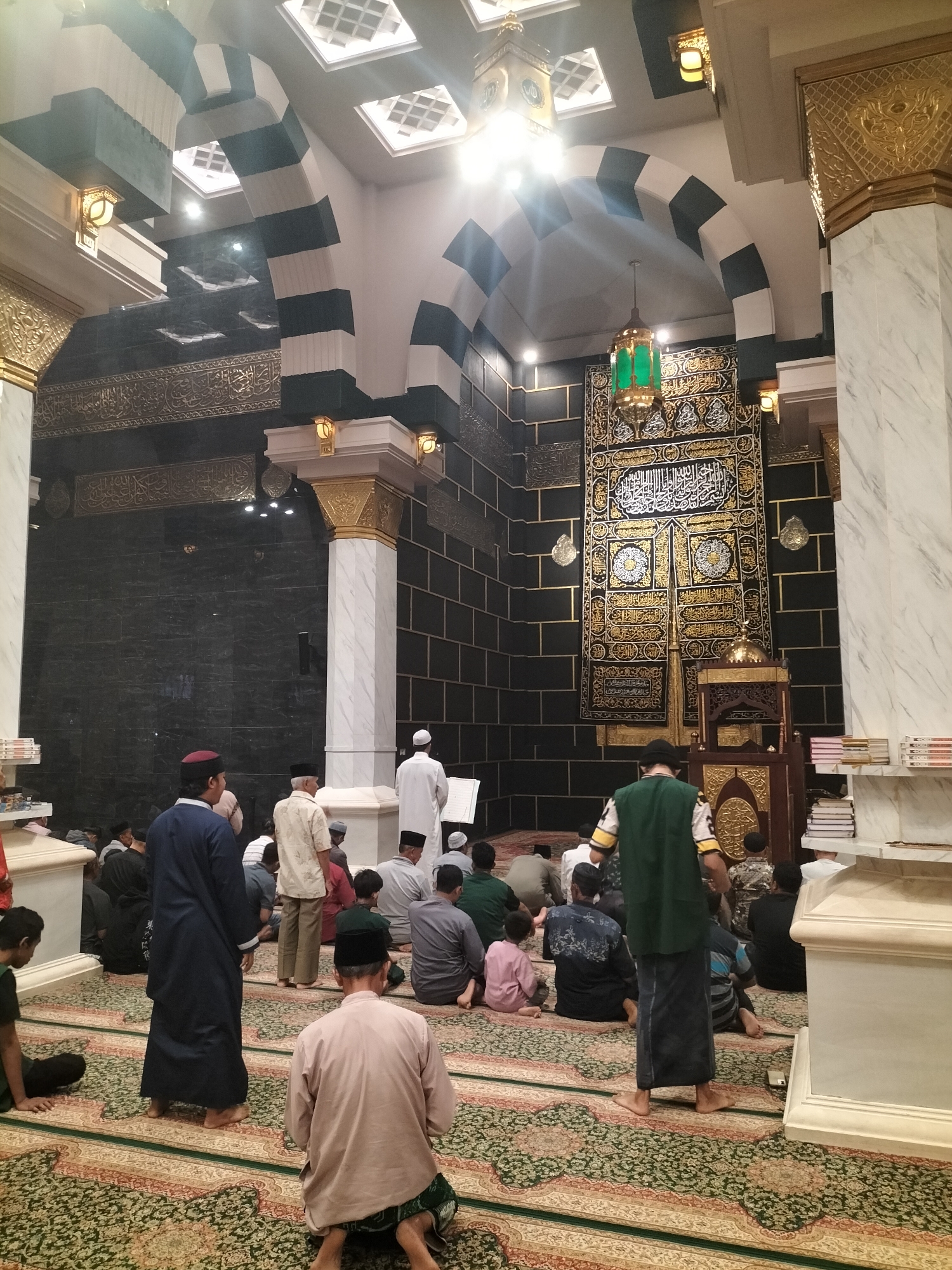The image shows the interior of a mosque, likely the Masjid Agung Sang Soopyan, located in Blitar, East Java, Indonesia. The mosque features distinctive architecture with arched doorways and patterned ceilings. The walls are adorned with Islamic calligraphy and decorative motifs. In the center of the mosque, a large, ornate tapestry with Arabic script hangs prominently. Several men are seated on the patterned carpet, facing towards the tapestry in prayer or contemplation. One man in a dark blue robe stands to the left, and another in a white garment stands to the right. The lighting suggests it is daytime, with natural light filtering in from above and artificial lights also illuminating the space. The overall atmosphere appears to be one of reverence and devotion.