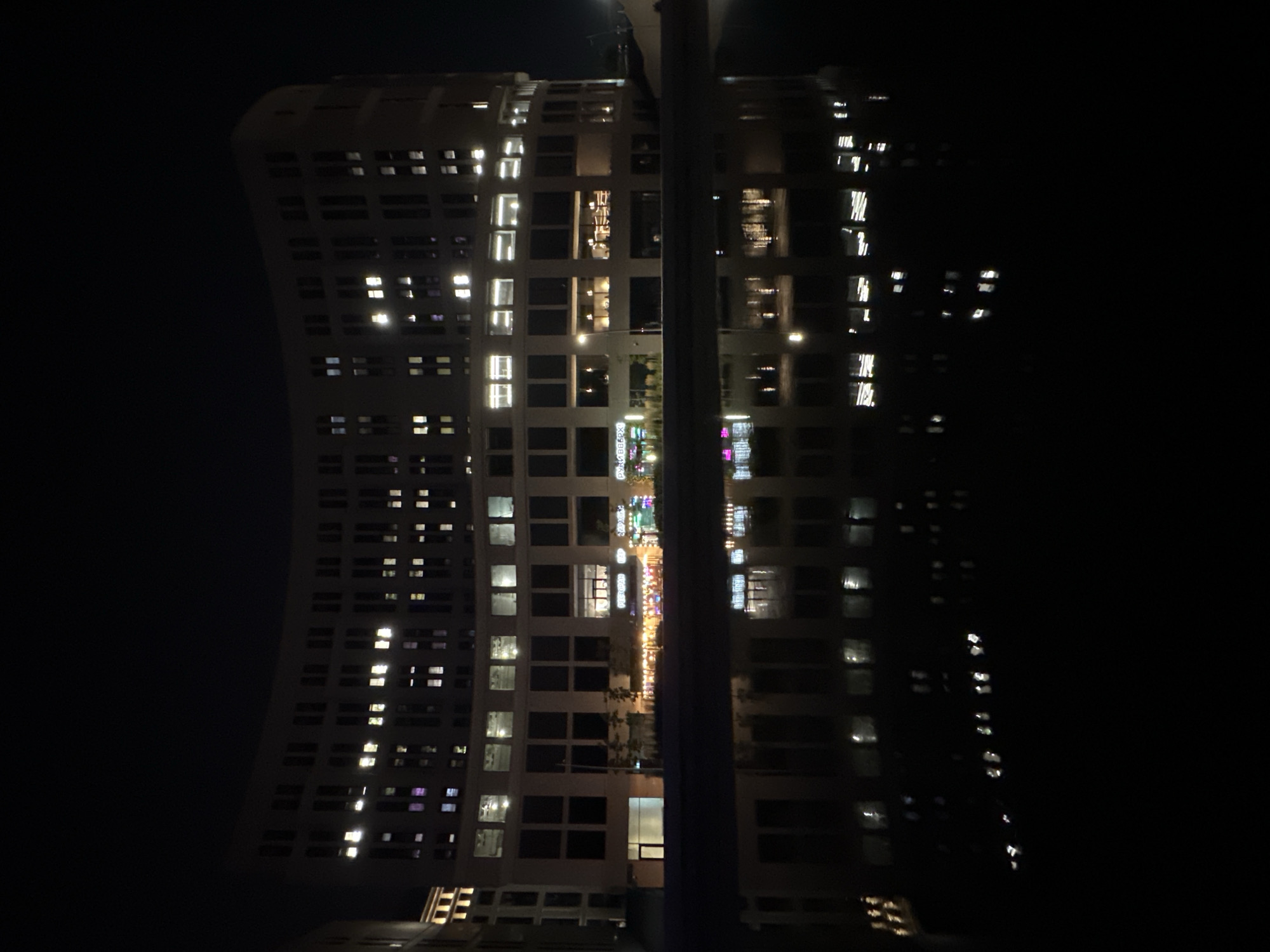 A nighttime, eye-level, medium shot captures a curved, multi-story building in the city of Unknown, Unknown, reflected in dark water. The building features numerous illuminated windows, creating a pattern of bright squares against the dark facade. The lower section of the building has a row of storefronts with visible lights and signage, though the text is illegible. Trees are present along the waterfront in front of the building. The reflection of the building and its lights is clearly visible in the still water below, creating a symmetrical, inverted image. The sky is entirely dark. There are no people or discernable actions captured in the scene.