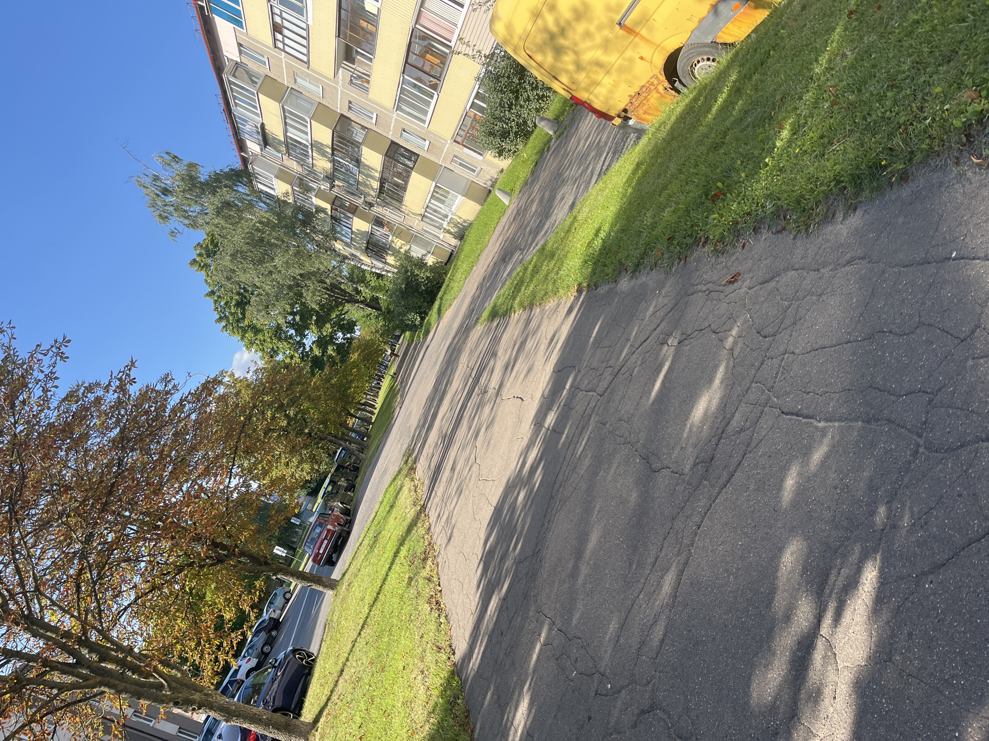 An outdoor, low-angle, medium shot captures a cracked asphalt pathway on the right, bordering a strip of green grass on the left. The pathway recedes into the distance, lined with parked cars. Further back, a multi-story, yellow apartment building with balconies stands on the right side of the frame, with trees and a bright blue sky behind it. On the left, trees with autumnal foliage are visible, casting shadows on the pathway and grass. The image appears to be taken in Minsk, Belarus, possibly during the autumn season.