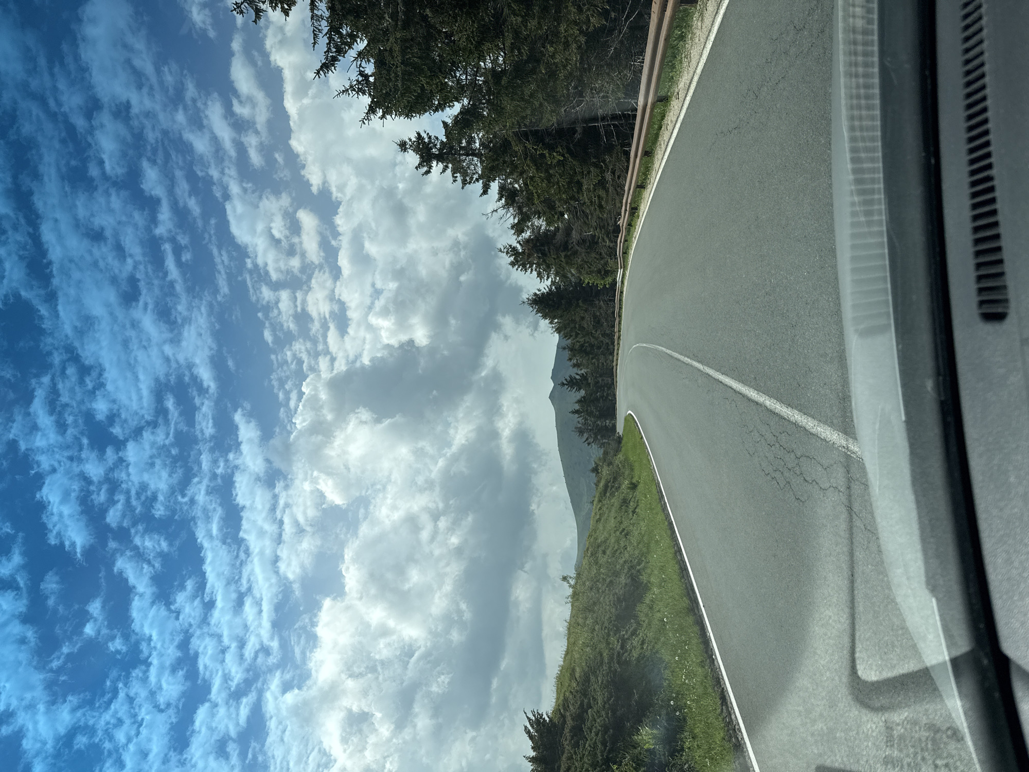 The image presents a forward-facing view from inside a vehicle traveling on an asphalt road in Hristo Danovo, Bulgaria. The road, featuring a solid white line on the left and a dashed white centerline, curves gently to the right and exhibits some surface cracking. To the left of the road, a verdant grassy embankment rises, dotted with green shrubs and small white flowers. To the right, a weathered brown metal guardrail parallels the road, backed by a dense line of dark green coniferous trees. In the mid-distance, rolling green hills or mountains are visible under a dynamic sky. The upper half of the frame is dominated by a bright blue sky filled with numerous white cumulus clouds, ranging from small, scattered formations at higher altitudes to larger, more substantial clouds closer to the horizon. The very bottom of the image shows the vehicle's dashboard and windshield, with reflections visible on the glass and a ventilation grate at the lowest edge. No individuals or animals are present.
