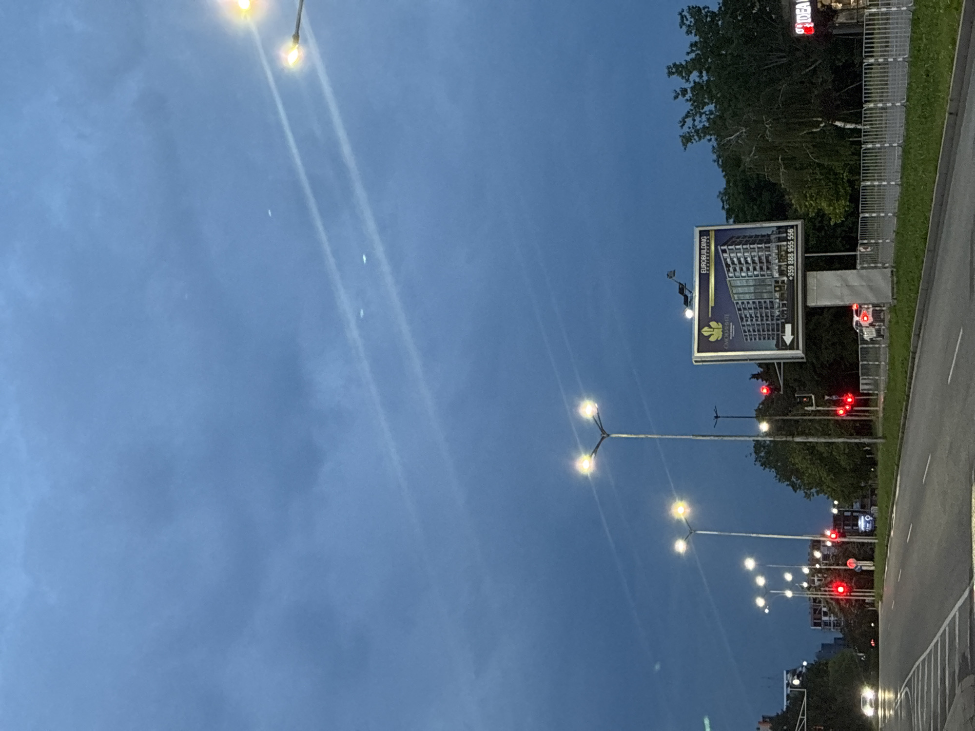 This image depicts an urban street scene during dusk or early evening, characterized by a clear, dark blue sky. Numerous streetlights are illuminated, casting bright, elongated streaks of light across the sky, possibly due to lens flare or atmospheric conditions.
On the right side of the frame, a multi-lane road is visible with distinct white markings. Several traffic lights glow red, suggesting vehicles are stopped or preparing to stop. Adjacent to the road, a large billboard prominently displays an advertisement for "EUROBUILDING," featuring an image of a modern high-rise building and a stylized leaf or plant logo. Below the building image, a phone number is visible: "+359 888 955 556." The country code +359 confirms the location in Bulgaria.
Behind the billboard and along the roadside, green trees and other vegetation are present. A metal fence or barrier runs parallel to the road. In the distance, other faint lights, including red glows, hint at additional urban infrastructure or buildings. The overall setting is a bustling city environment at the transition from day to night, with no specific people or direct activities clearly visible, but implying ongoing traffic and city life.