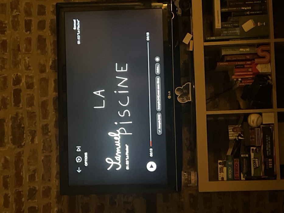 In a dimly lit interior setting in Carnin, France, a black LG flat-screen television is centered against a light brown brick wall with dark mortar. The television displays a streaming service interface with a black background.

On the screen, the title "LA Samuel Piscine" is prominently shown in white text, with "LA" in block letters and "Samuel Piscine" in a script-like font. Above the title, in the top right corner, "Samuel" and "S1: E3 'La Piscine'" are displayed. In the top left, navigation icons (back, reload/restart, fast forward) and the word "OPTIONS" are visible. At the bottom of the screen, a play button is present next to the current playback time of "00:13". A red progress bar extends to the right, with the total duration "04:18" marked at its end. Audio and subtitle options are visible, with "français [VO]" currently selected and "français [VO] avec sous-titres" and "Autre..." as further choices.

Below the television, on a light-colored shelving unit, several objects are arranged. Directly beneath the screen, from left to right, a clear glass object (possibly a candle holder) with a label is present. Centered on the TV's base is a black silhouette cutout of two embracing figures, with a photograph inserted. To the right of the silhouette, two small, light-colored rectangular objects are positioned next to a vintage-style camera with a silver lens.

The shelving unit consists of at least two visible cubbies. The left cubby contains multiple stacked board games or boxed items, including "SKYJO," "Qwirkle," "CODE NAMES," and "GRIMM." A small baseball is visible on the right side of this cubby. The right cubby holds a collection of books with various spine colors, predominantly red and orange, arranged vertically. A prominent red letter "S" sculpture or ornament stands among the books.