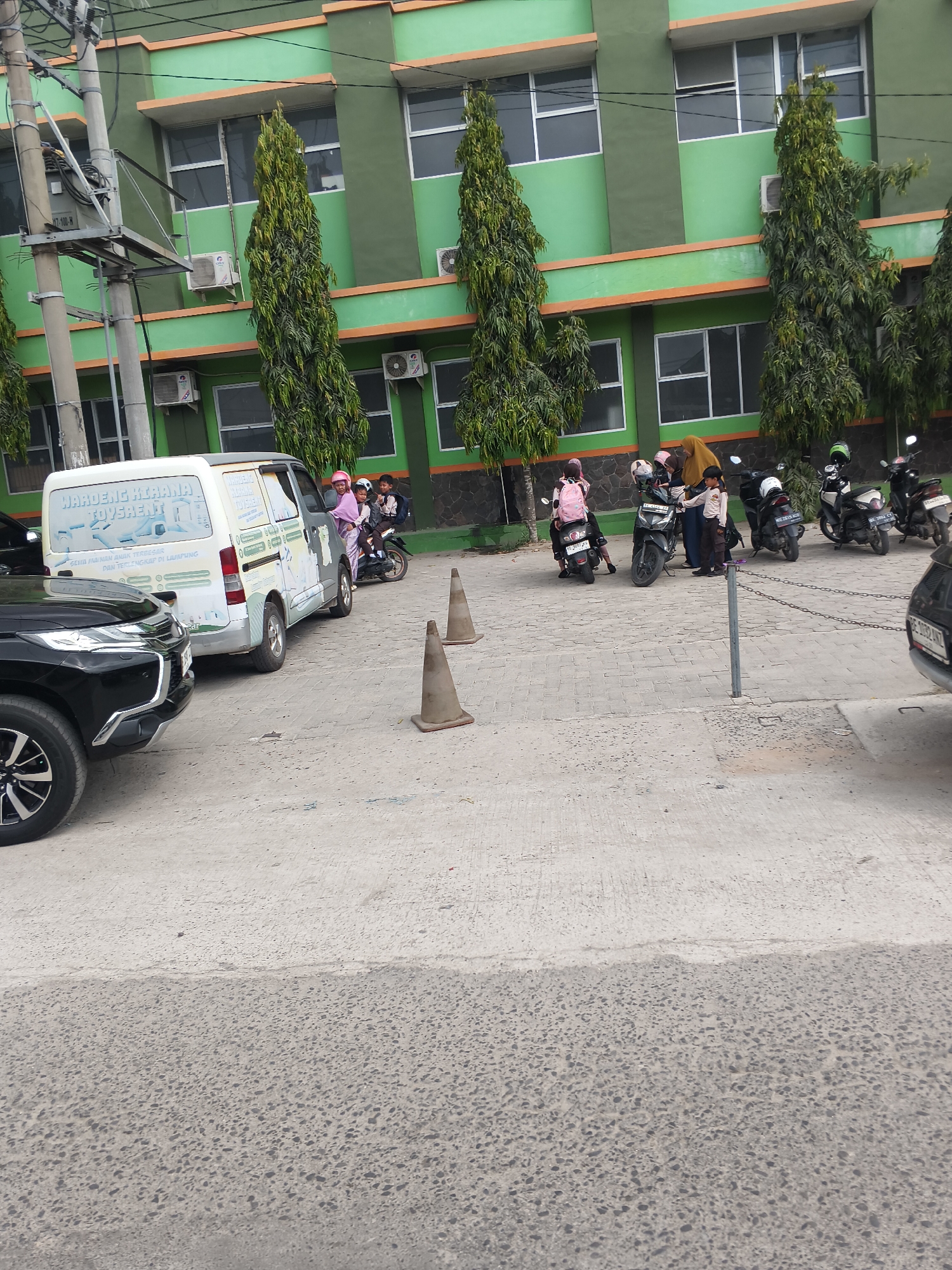 This is a street-level, medium shot photograph taken in Bandar Lampung, Indonesia, capturing a scene in front of a building with a distinctive green facade and orange trim. The building appears to be a school or similar institution, evidenced by the presence of children and adults with what seem to be school bags and uniforms.
In the foreground, a black SUV is parked on the left side of the frame, with its front passenger side wheel and bumper visible. Next to it, a white van with decals and text that appears to advertise a service is parked. Several motorcycles are lined up in the middle ground, with a few individuals, including children, either on or near them. One child, wearing a pink headscarf and backpack, is seated on the back of a motorcycle. Another child, in a school uniform, is standing and appears to be interacting with an adult. A woman in a yellow headscarf is also visible.
Two orange traffic cones are placed on the paved ground in front of the van. In the background, the building's windows are visible, and several tall, slender evergreen trees are planted along the building's perimeter. A chain-link fence with metal posts runs across the middle ground, separating the parking area from a walkway or another section of the grounds. Electrical poles and wiring are visible to the far left of the frame, indicating urban infrastructure. The overall lighting suggests it is daytime, likely under overcast or diffused sunlight conditions.