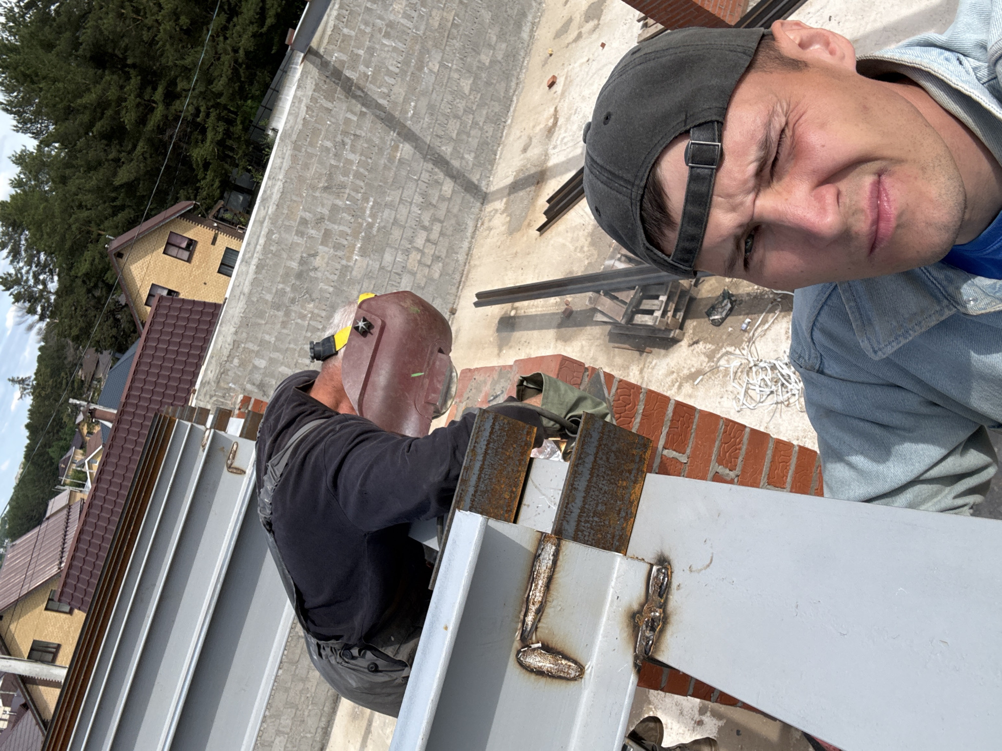 This media file captures a selfie taken on what appears to be an elevated construction site or rooftop in Chaikovsky, Russia, during the daytime under clear, sunny weather.
The primary subjects are two men. On the left side of the frame, a man is engaged in welding. He is seen wearing a dark brown welding helmet with a yellow accent, a black long-sleeved shirt, and dark work pants or overalls. His face is obscured by the helmet, and he is wearing work gloves. He is positioned next to partially welded metal beams, which show visible, somewhat uneven weld marks. The metal structures are a light gray color, with some parts showing rust.
The second man, positioned closer to the viewer on the right, is taking the selfie. He is wearing a dark gray baseball cap backward, a light blue denim jacket over a blue t-shirt. His expression appears squinted or strained, possibly due to the bright sunlight or the effort of taking the photo.
The setting includes a mix of construction elements and a residential landscape. Behind the welder, there are sections of brick wall and concrete flooring. Various metal beams and construction debris are scattered on the ground. In the background, beyond the construction area, several residential houses with brown and yellow tiled roofs are visible. A dense line of green trees, possibly conifers, forms the horizon, suggesting a semi-rural or suburban environment. The angle of the shot indicates a significant height, looking down upon the surrounding area. There is no visible text in the image.