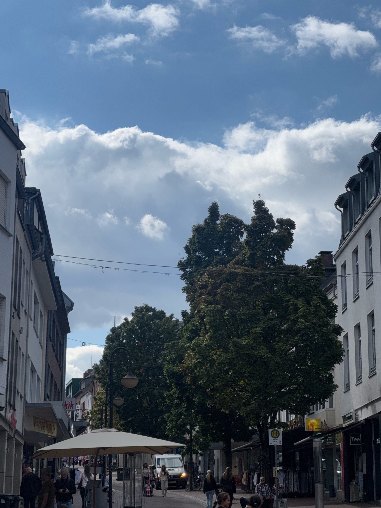 The image depicts a street scene in Cleves, Germany, featuring buildings lining both sides of a pedestrian walkway. The sky above is partly cloudy with a bright blue sky visible in patches and large, puffy white clouds dominating the upper portion of the frame.
On the left side of the street, a white building with multiple windows and a gray roof can be seen. Signs for "Kik" and "Playboy Fantasy" are visible on the storefronts. People are walking on the sidewalk.
On the right side of the street, a white building with dormer windows and gray roofs is prominent. A sign with the letter "H" in a yellow circle, indicating public transport, is mounted on a pole. Storefronts with awnings are visible, displaying clothing racks outside. One sign reads "Tee."
In the center of the image, large, leafy green trees with dark green foliage are the most striking natural elements. A street lamp with two lights is positioned between the trees and the right-hand buildings. A large, light-colored umbrella is set up near the foreground, possibly for an outdoor seating area. A white van is parked on the street.
Several individuals are present in the scene, walking along the street and sidewalk. Their clothing suggests a casual daytime setting. The overall atmosphere is that of a commercial street in a European town.