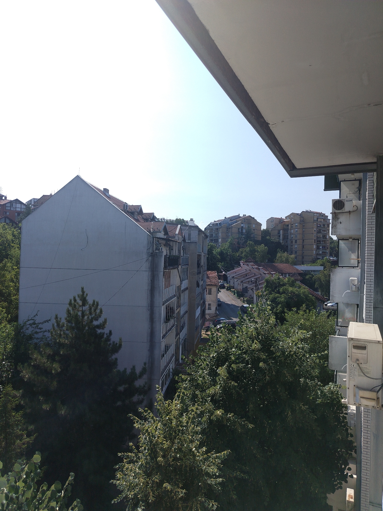 The image captures a view from a balcony looking out over a residential area in Cukarica Urban Municipality, Serbia. In the foreground, a portion of the balcony ceiling and a building facade with air conditioning units are visible. The main focus is on the urban landscape, with several apartment buildings of varying heights and architectural styles dominating the scene. Some buildings are clad in lighter-colored materials, while others have a more aged appearance. A street can be seen winding through the buildings, with parked cars suggesting a typical urban setting. Lush green trees and foliage are interspersed among the buildings, indicating a relatively well-treed neighborhood. The sky is clear and bright, suggesting a sunny day. No people are clearly visible in the image.