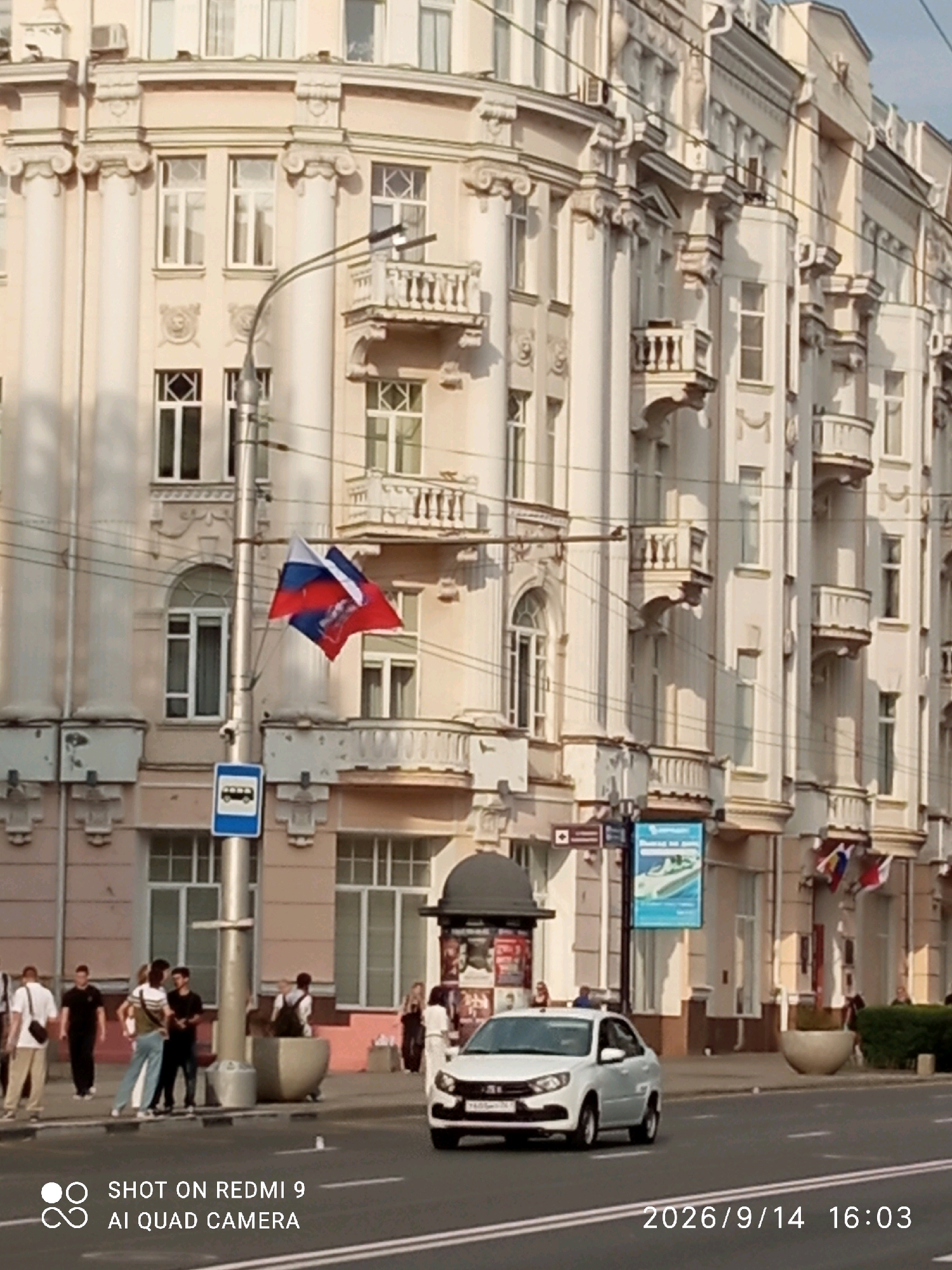 A street-level, eye-level, medium shot captures a city street scene in Rostov-on-Don, Russia. The main subject is a pale pink, ornate, multi-story building with classical architectural elements, including columns, balconies with railings, and decorative carvings. A street lamp with two flags, one appearing to be the Russian flag, is positioned in front of the building.
In the foreground, a white sedan drives on the road. A bus stop sign is visible near the street lamp. Several people are walking on the sidewalk or standing near the bus stop. The environment appears to be daytime, with sunlight casting shadows. The road has white lane markings. The lower right corner of the image displays a timestamp and camera information: "2026/9/14 16:03" and "SHOT ON REDMI 9 AI QUAD CAMERA".