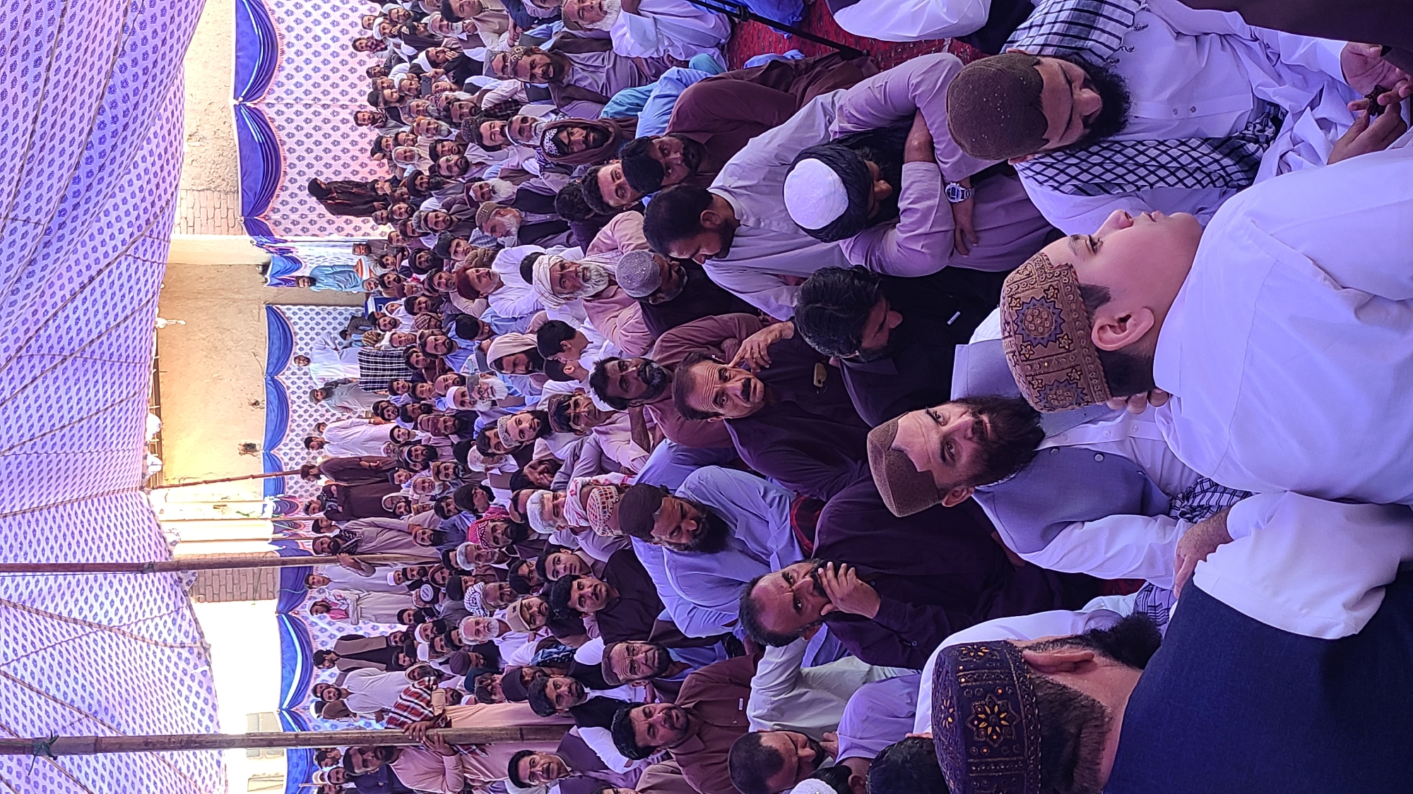 A wide, medium-angle shot captures a large gathering of men sitting under a tent in Panjpai, Pakistan. The tent is supported by wooden poles and adorned with a patterned, blue and white fabric. The backdrop includes a tan wall and visible portions of other structures. The men in the crowd are primarily dressed in traditional Pakistani clothing, with many wearing white or light-colored kurtas and a variety of head coverings, including kufis and turbans. Their expressions vary from attentive to contemplative, and some appear to be engaged in conversation. The overall atmosphere suggests a formal or religious event, given the attire and the arranged seating. The photograph seems to be taken from a vantage point that provides a clear view of a significant portion of the gathering.