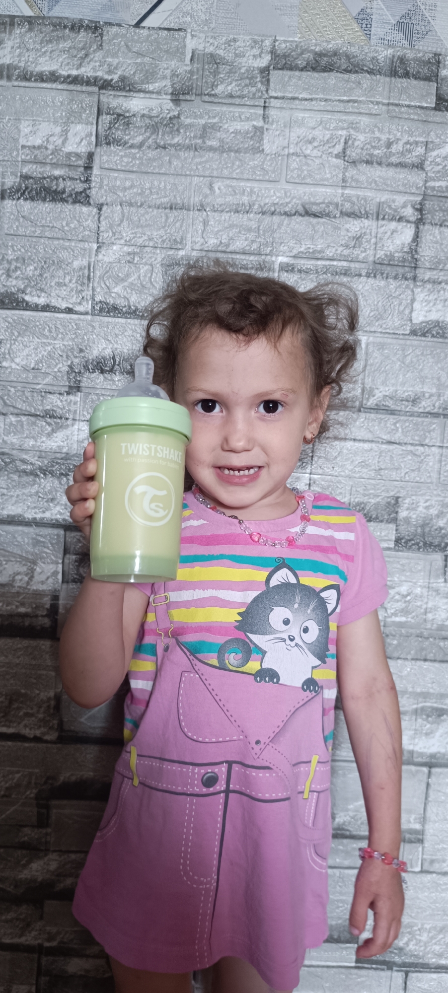 In an indoor setting with a textured gray brick-patterned wall as the background, a young girl stands holding a light green baby bottle. She is wearing a pink shirt with horizontal stripes of yellow, blue, and white, layered with a pink overall-style garment featuring a graphic of a cartoon cat peeking out of a pocket. A pink beaded necklace adorns her neck, and a pink and clear beaded bracelet is on her right wrist. The girl is looking directly at the camera and smiling, displaying her teeth. The baby bottle, held in her right hand, is partially filled with a light yellow liquid and has "TWISTSHAKE" printed on it. The image does not provide any information about the city of Karsha, Russia.