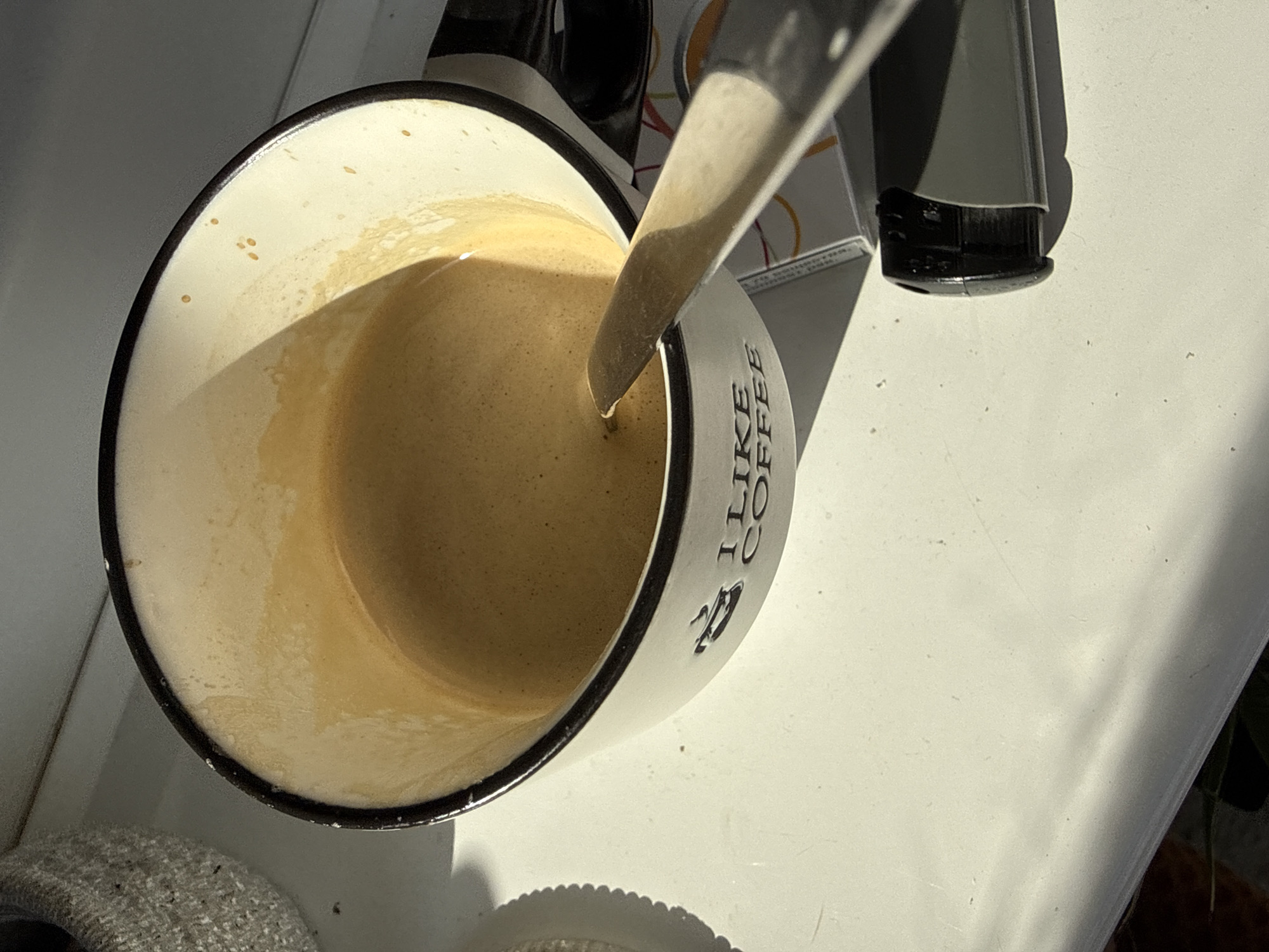 An overhead shot, slightly tilted, captures a mug of coffee or espresso on a white surface, likely a countertop or table, in Dobrogled, Bulgaria. The mug is white with a dark rim and features the text "I LIKE COFFEE" printed vertically on its side, accompanied by a small graphic resembling a coffee cup. A spoon is inserted into the amber-colored beverage, which has a layer of crema on top.
Bright, direct natural light, indicative of a sunny day, illuminates the scene from the right, casting distinct shadows from the mug, spoon, and an object behind the mug. This object appears to be part of a coffee machine or a similar appliance, with dark grey and black components visible.
To the bottom left, a textured, grey fabric object, possibly a coaster or part of a towel, is partially visible. The white surface exhibits some minor imperfections and specks. The overall impression is one of a freshly prepared coffee beverage, possibly being stirred, bathed in warm sunlight.