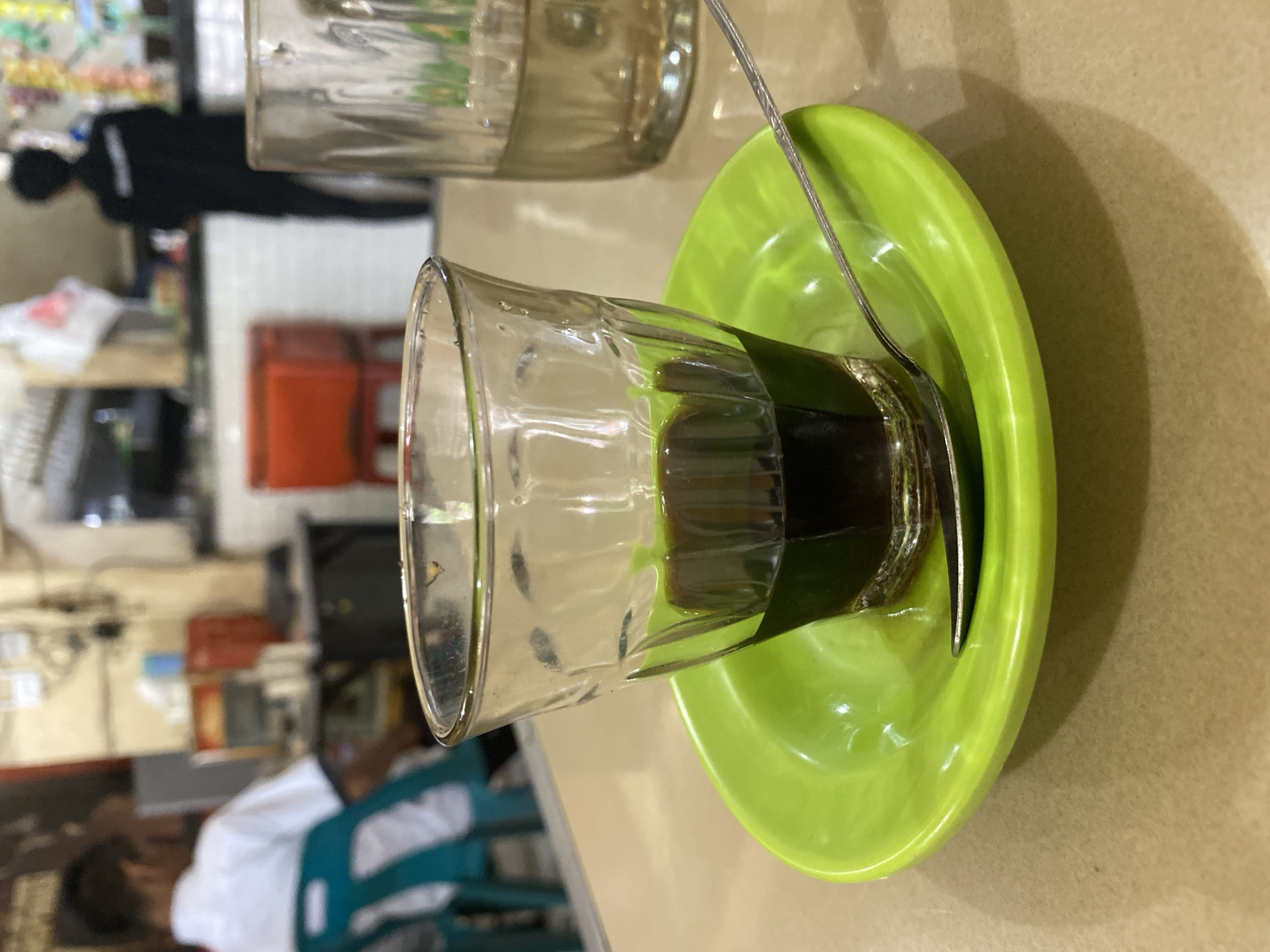A close-up shot features a small glass filled with dark liquid, likely coffee, resting on a vibrant green saucer. A silver spoon is placed on the saucer. In the background, the scene depicts a busy cafe or restaurant environment. To the left, a person in a white shirt is seated facing away from the camera. Further back, another individual in a dark uniform stands behind a counter. The cafe appears to be located in Lhokseumawe, Indonesia, suggested by the overall ambiance and style of the establishment. The lighting is warm and slightly dim, creating a cozy atmosphere.