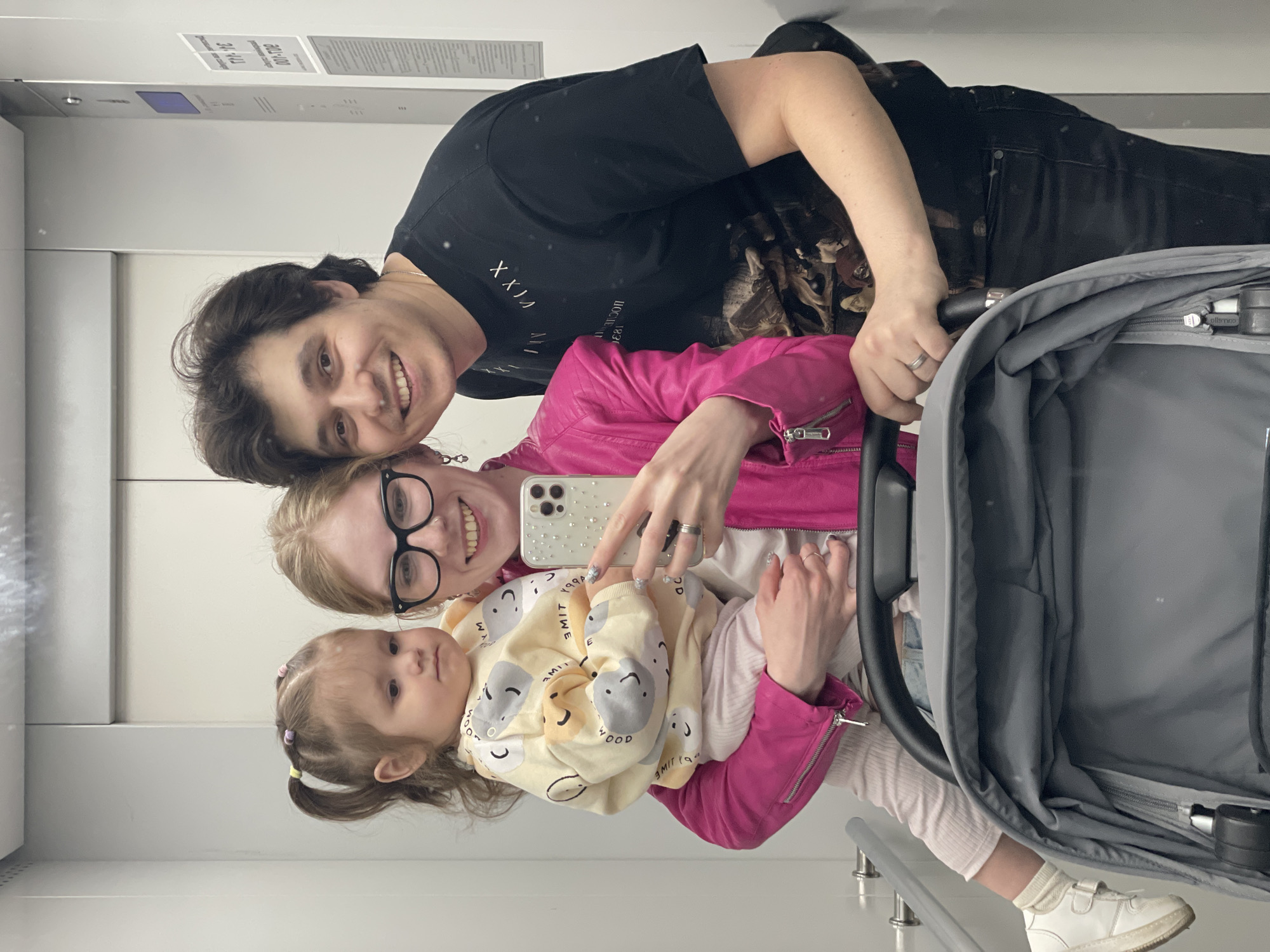 The image is a selfie taken by a woman with glasses, wearing a pink jacket, holding a phone with a patterned case. She is standing in what appears to be an elevator, as indicated by the signage in the background. She is holding a small child in a patterned sweater. A man in a black t-shirt with text is visible behind the woman. A stroller with a gray interior is in the foreground. The text in the image suggests that this is in Izhevsk, Russia.