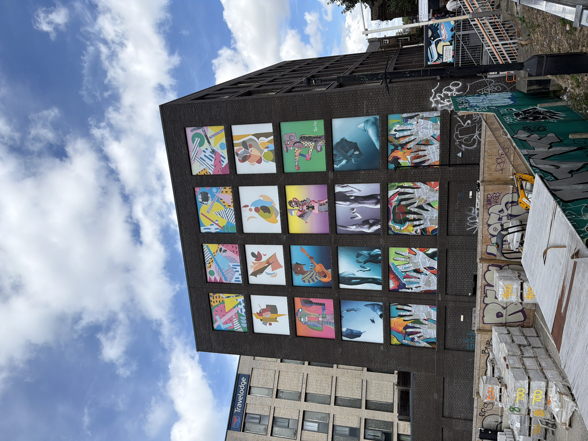 This image captures a building in London, United Kingdom, adorned with a grid of colorful artworks on its dark brick facade. The artworks are square and arranged in four rows and three columns. In the background, a Travelodge hotel building is visible to the left. In the foreground, construction materials, including stacks of pallets wrapped in plastic and covered with yellow markings, are present. Graffiti is visible on a wooden fence and a green barrier. A lamppost stands to the right of the building, and a set of stairs leads up to a platform. The sky is partly cloudy with blue patches.