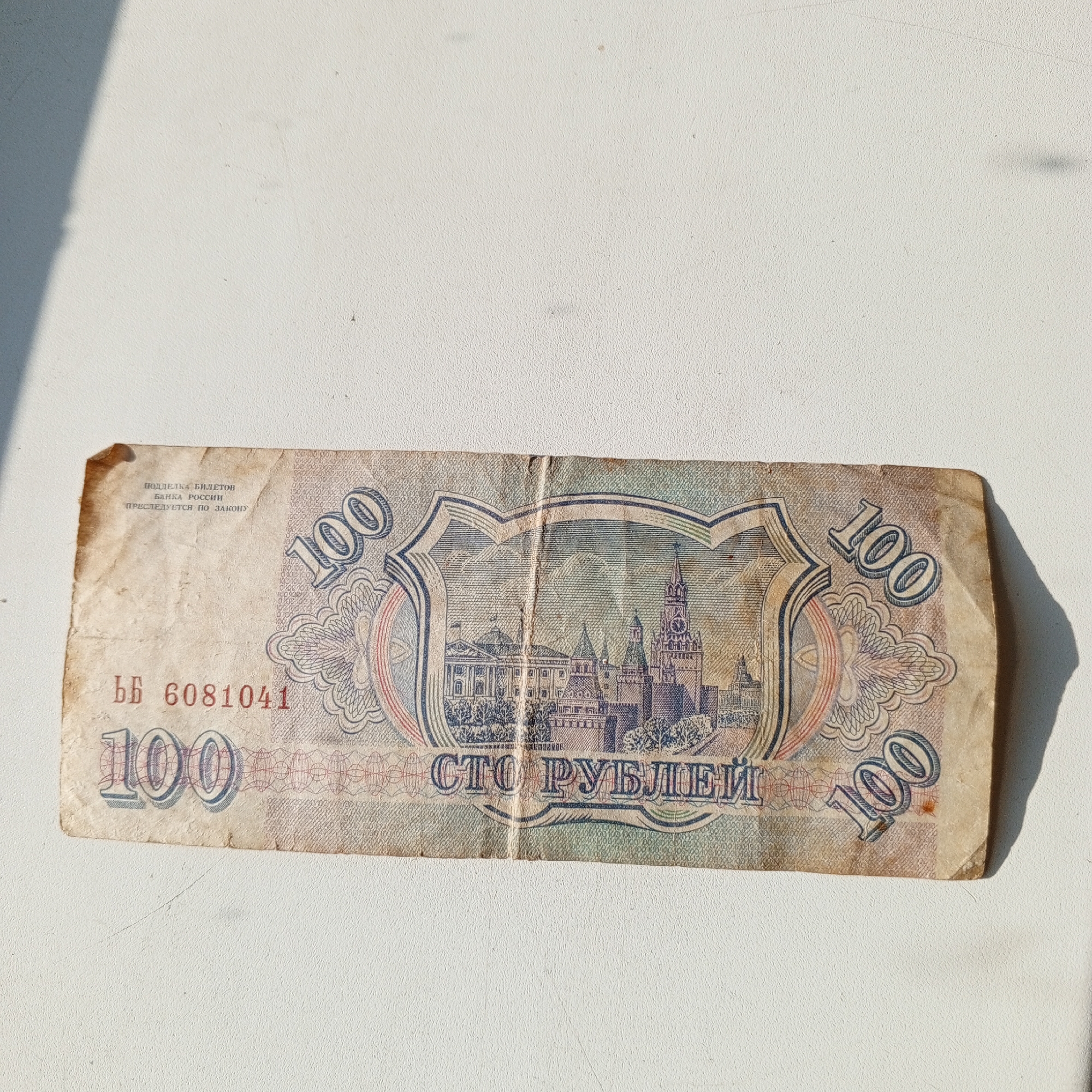 The image shows a single, folded, aged banknote lying on a white, textured surface. The banknote is predominantly light brown and beige with some blue and red accents. The front of the banknote is visible, displaying an ornate design with Arabic numerals "100" prominently featured on the left and right sides. In the center, a detailed illustration depicts architectural structures, including what appears to be a Kremlin-like fortress with towers and a dome. Text in Russian is also visible, including the phrase "СТО РУБЛЕЙ" (STO RUBLEY) which translates to "ONE HUNDRED RUBLES." A serial number "Bb 6081041" is printed on the lower left side. The banknote is creased and shows signs of wear. The white surface appears to be a wall or tabletop, with a shadow cast by an unseen object on the left side of the image. The environment is brightly lit, suggesting outdoor lighting or a well-lit indoor space. There is no indication of the city of Sayanogorsk, Russia, or any specific location within it.