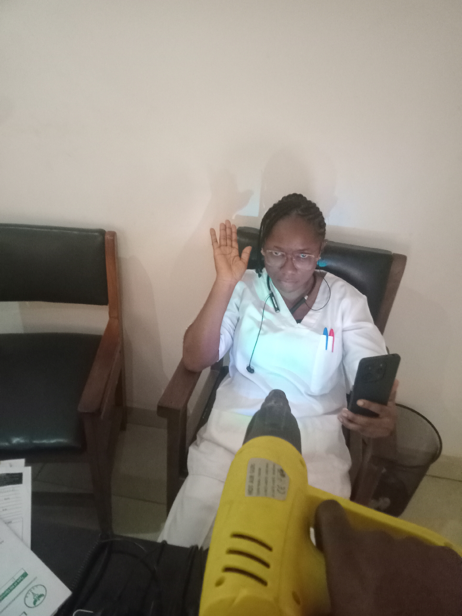 In an indoor setting in Gwarinpa, Nigeria, a woman in a white medical uniform is seated in a dark, high-backed chair with wooden arms. She appears to be a healthcare professional, wearing glasses and a headset around her neck, with her hair styled in braids. Two pens, one red and one blue, are clipped to her uniform pocket. Her right hand is raised, palm facing forward, as if in a greeting, pledge, or a gesture of acknowledgement. In her left hand, she holds a black smartphone, which she is looking at, or is positioned for a video call or recording.
The background consists of light-colored, plain walls. To her left, another dark chair with wooden arms is visible. On a surface to the left foreground, several papers are stacked, one showing a green circular logo that resembles a tree or plant. A small, black, cylindrical bin is located near her left arm.
In the immediate foreground, a yellow tool, possibly a heat gun or a hair dryer, is held by a person whose dark-skinned hand is visible, pointing towards the woman. The tool has an unreadable white label with black text on its side. The lighting in the room is predominantly artificial, casting a subtle blueish hue on the woman's uniform, and her expression is neutral to slightly serious. The scene suggests an interaction or presentation, possibly involving the phone and the person holding the yellow tool.