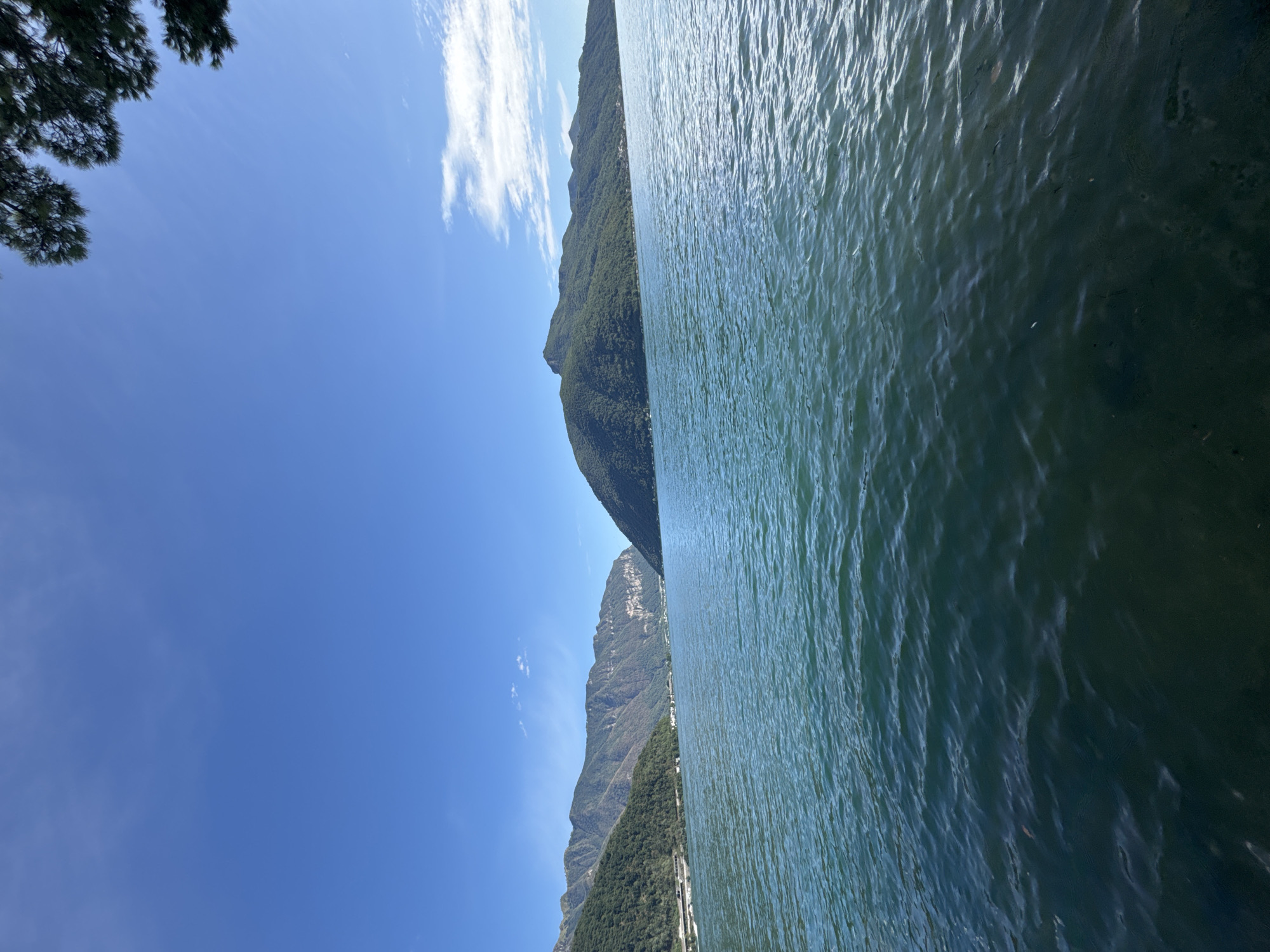 The image depicts a wide expanse of deep blue water, likely a lake, under a clear blue sky with a few scattered white clouds. In the background, lush green mountains rise dramatically from the water's edge, their slopes covered in dense vegetation. A road or highway can be seen winding along the base of the mountains on the left side of the image. The overall scene suggests a serene and natural landscape, possibly a popular tourist destination. The perspective is from water level, looking towards the mountains and the horizon. The presence of a tree branch in the upper right corner indicates that the photo was taken from the shore or from a boat close to the shore. Given the description of Melide, Switzerland, this scene likely captures a view of Lake Lugano.