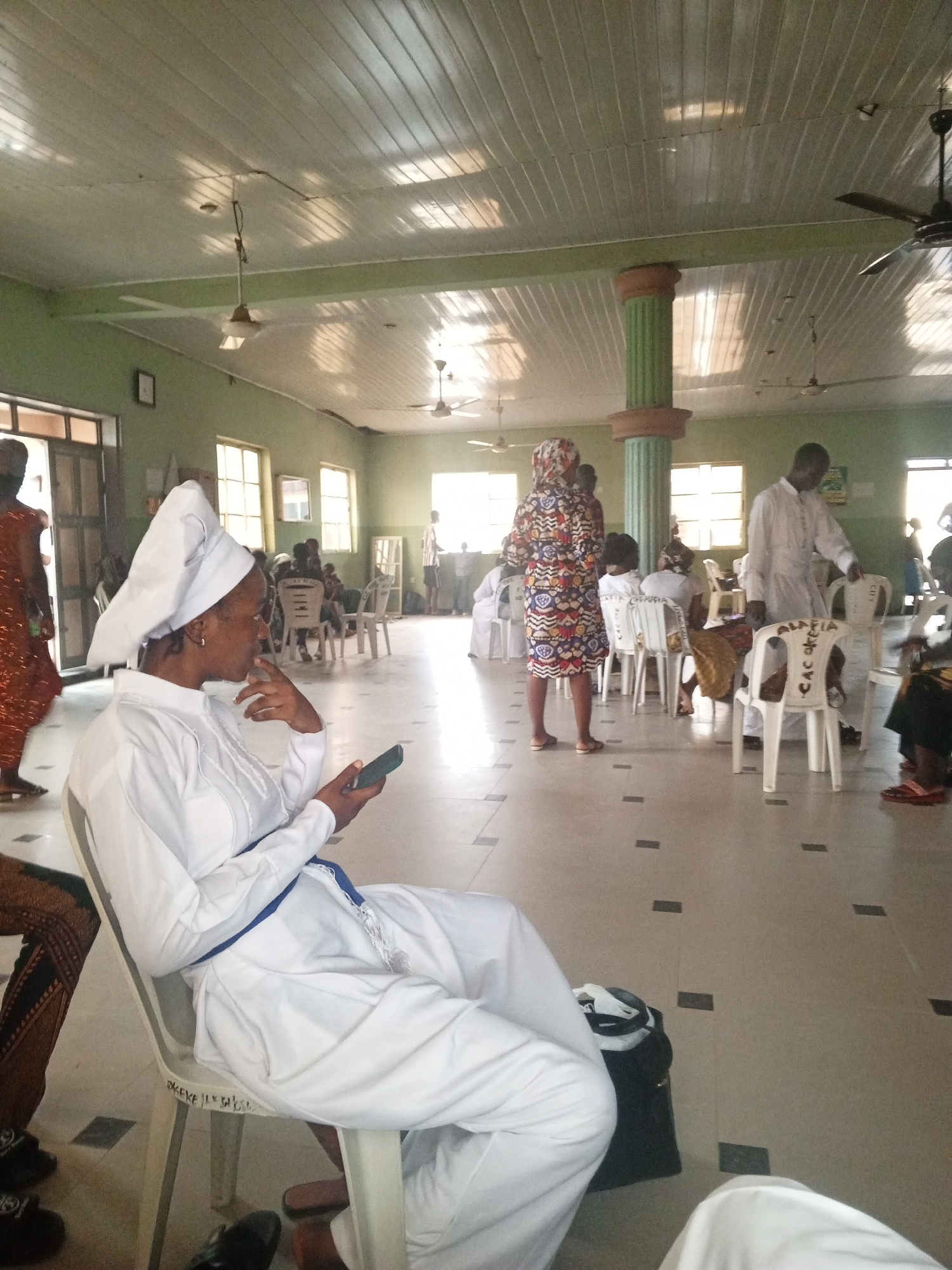 The image shows a group of people in what appears to be a large, indoor space. The room is painted with light green walls and has a white, panelled ceiling with ceiling fans. There are several windows providing natural light. Many individuals are seated on white plastic chairs arranged in rows. One woman in the foreground, dressed in white clothing with a white head covering, is seated and looking at a mobile phone. Others are wearing a variety of clothing, including a man in white robes. The floor is tiled with a light colour and accented with darker rectangular inlays. There is a large pillar in the room with green and brown accents.