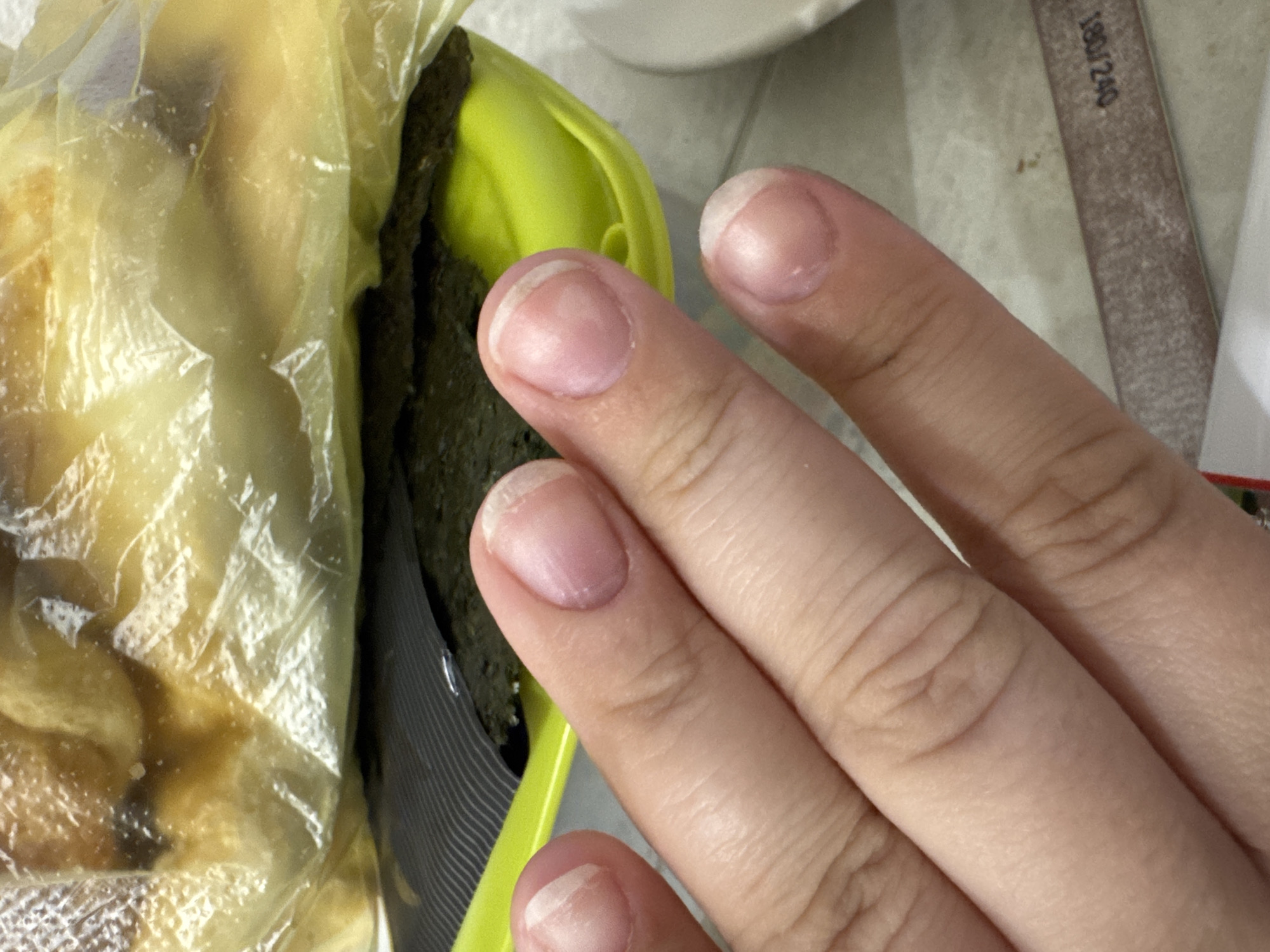 The image shows a close-up view of a person's hand with unpolished fingernails, resting on a surface that appears to be a countertop. In the background, a bright green plastic container holds dark green seaweed sheets, partially covered by a clear plastic bag containing food items. A nail file is visible on the countertop in the lower right portion of the image, with the grit rating "180/240" printed on it. The lighting suggests an indoor setting. There is no indication of the specific location within the city of Saratov, Russia.