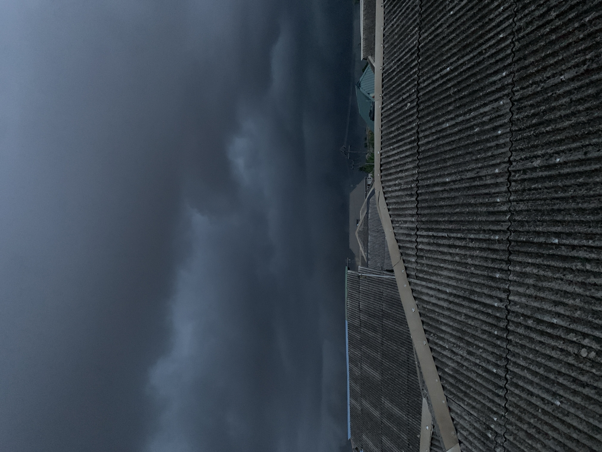 The image shows a dark, ominous sky over a building in Ikorodu, Nigeria. The sky is filled with dark, heavy clouds. A corrugated roof dominates the foreground, likely a part of a building or structure. In the middle ground, glimpses of buildings and possibly a power line are visible. The overall environment suggests a stormy or overcast day.